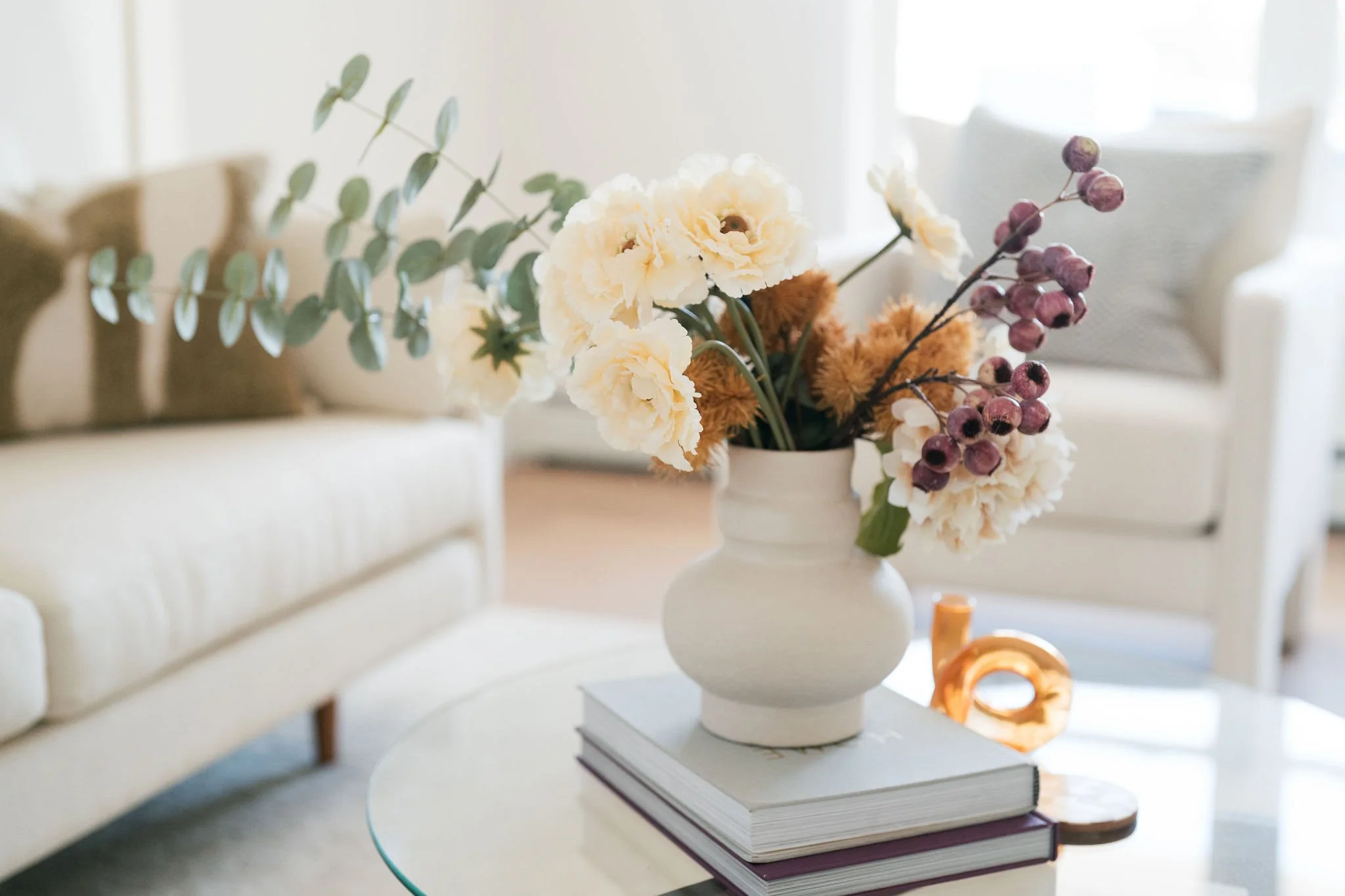 A white vase with a bouquet of flowers including cream-colored carnations, purple berries, and green leaves on a stack of books on a white table in a bright room.