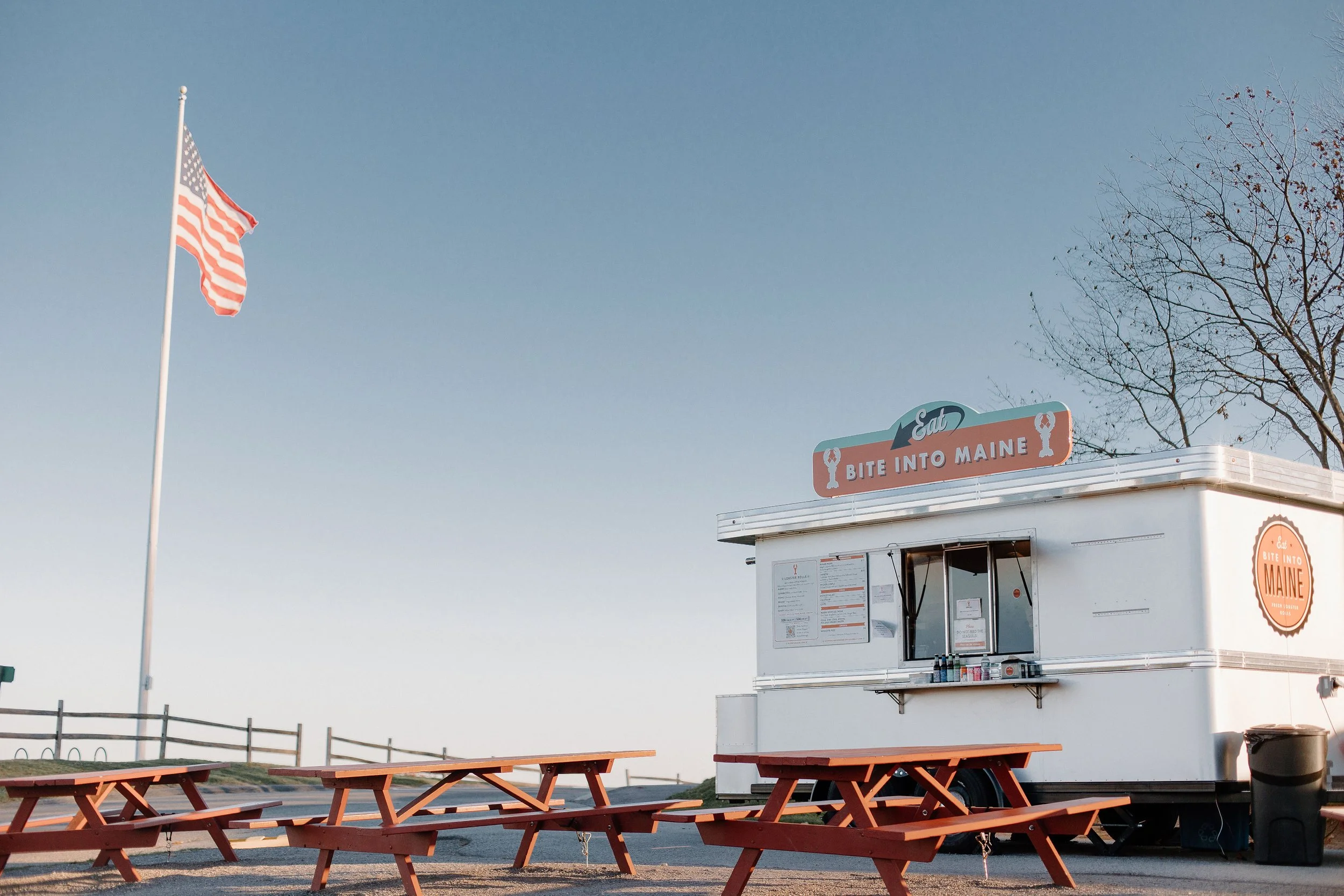 A food truck with a sign that says "Eat Bite Into Maine" under a clear blue sky; an American flag on a pole and several orange picnic tables in the outdoor seating area, with a leafless tree in the background.