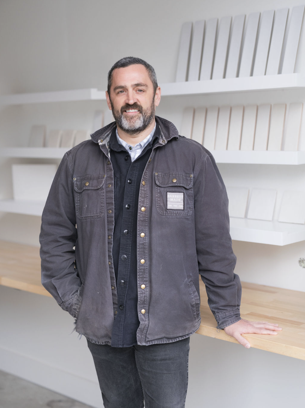 A man with a beard and short dark hair, wearing a black jacket over a button-up shirt, standing in front of white shelves with wooden samples in a bright, modern space.