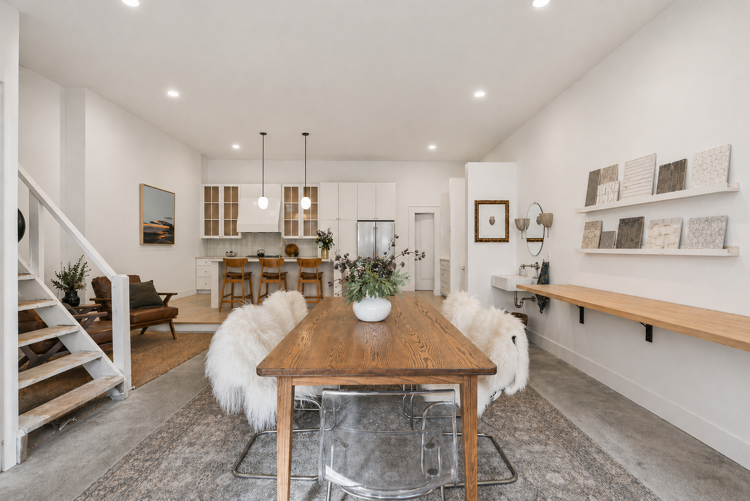 Open-concept dining and kitchen area with a wooden dining table surrounded by furry white chairs, a vase with flowers on the table, a kitchen counter with barstools, white cabinetry, and decorative tiles on the wall.