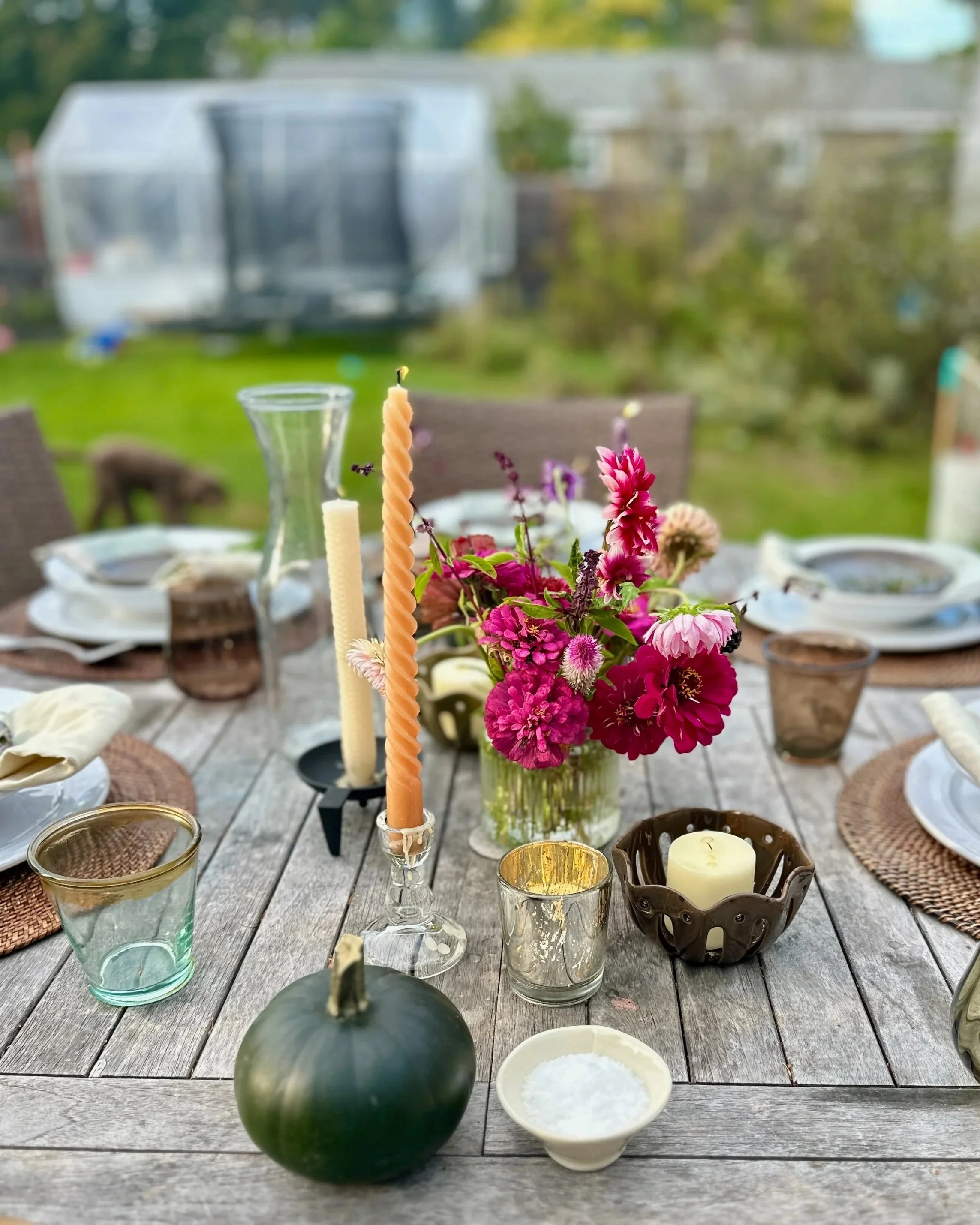 A close-up of a table setting outdoors with a bouquet of pink and purple flowers, candles, glasses, and plates on a wooden table.