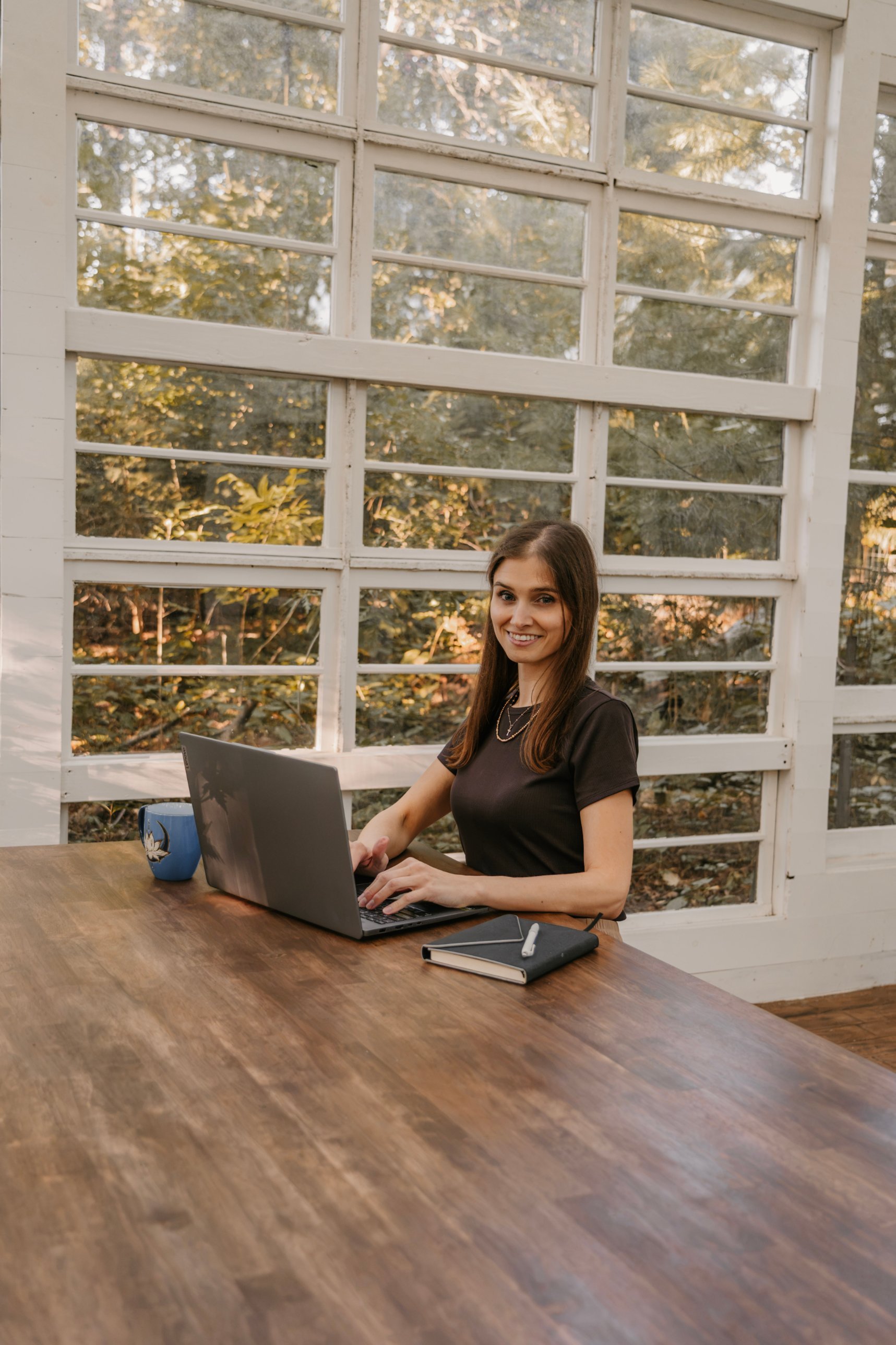 Cassandra working on a laptop at a wooden table by a sunny window