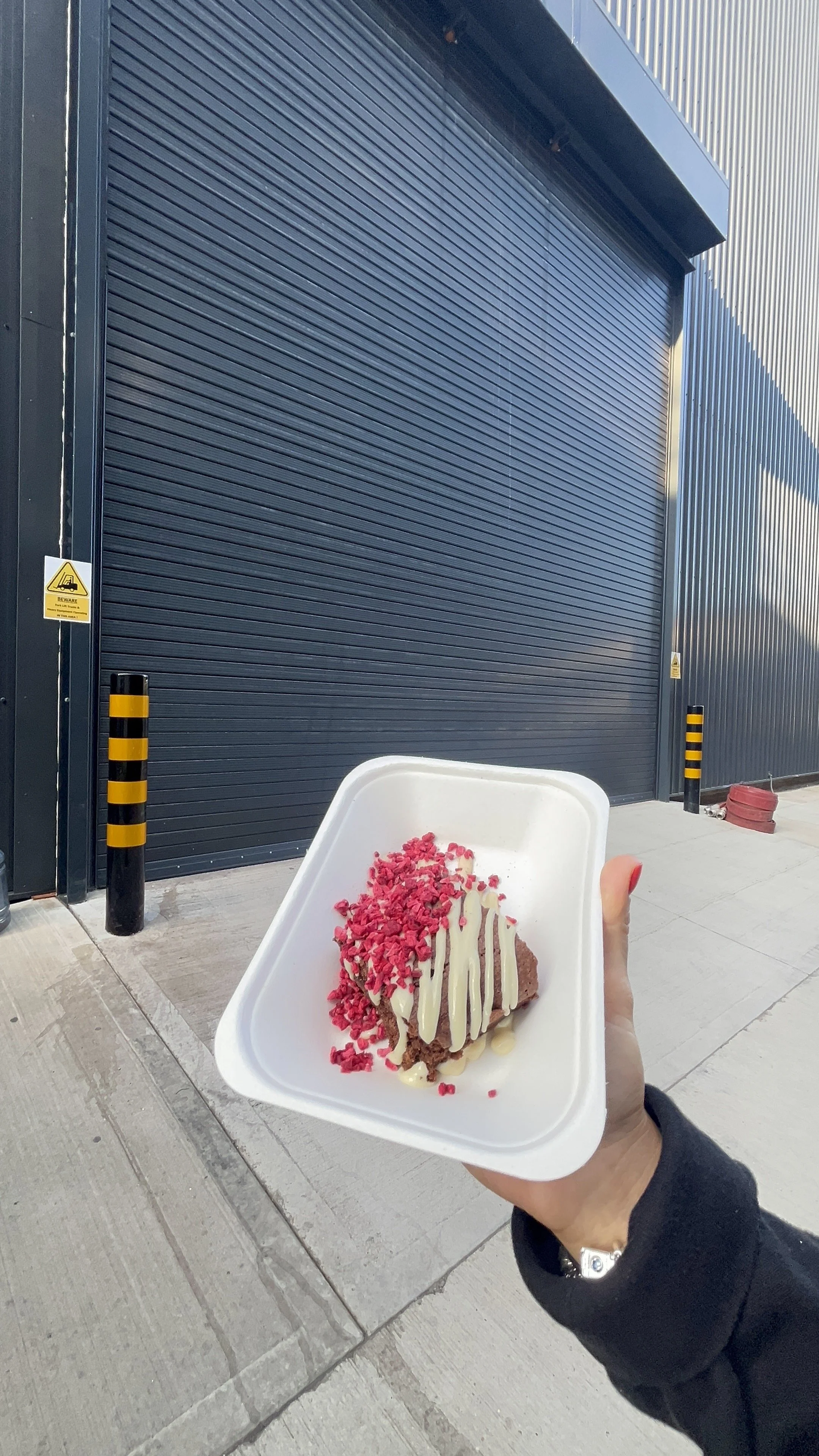 Person holding a white tray with a slice of chocolate cake topped with pink sprinkles and white chocolate drizzle, against a background of a closed dark garage door and sidewalk.