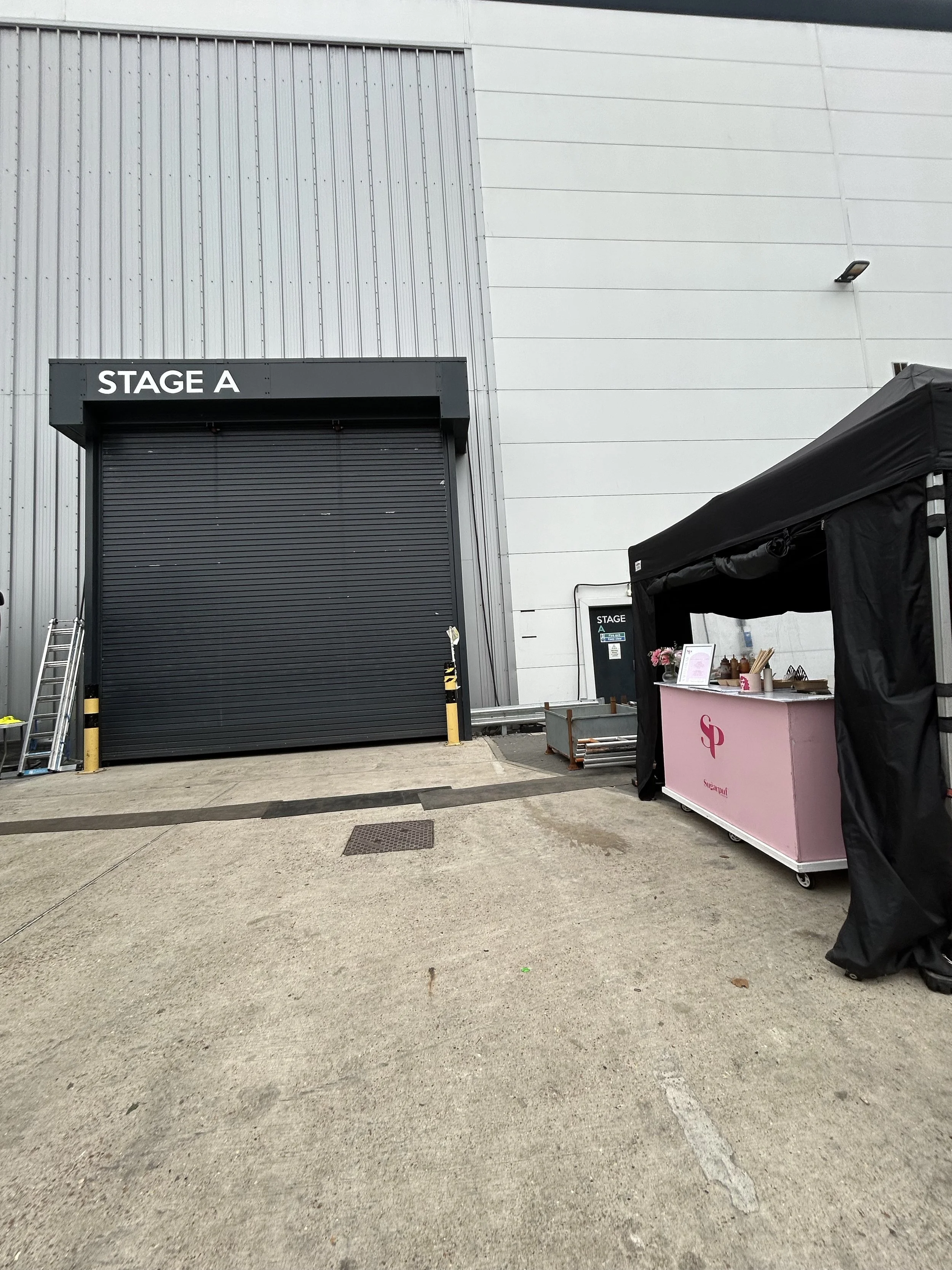 Exterior of a building with a large black door labeled 'STAGE A' and a small pink counter area on the right side under a black canopy. There are ladders and some equipment near the door, and the ground is concrete.