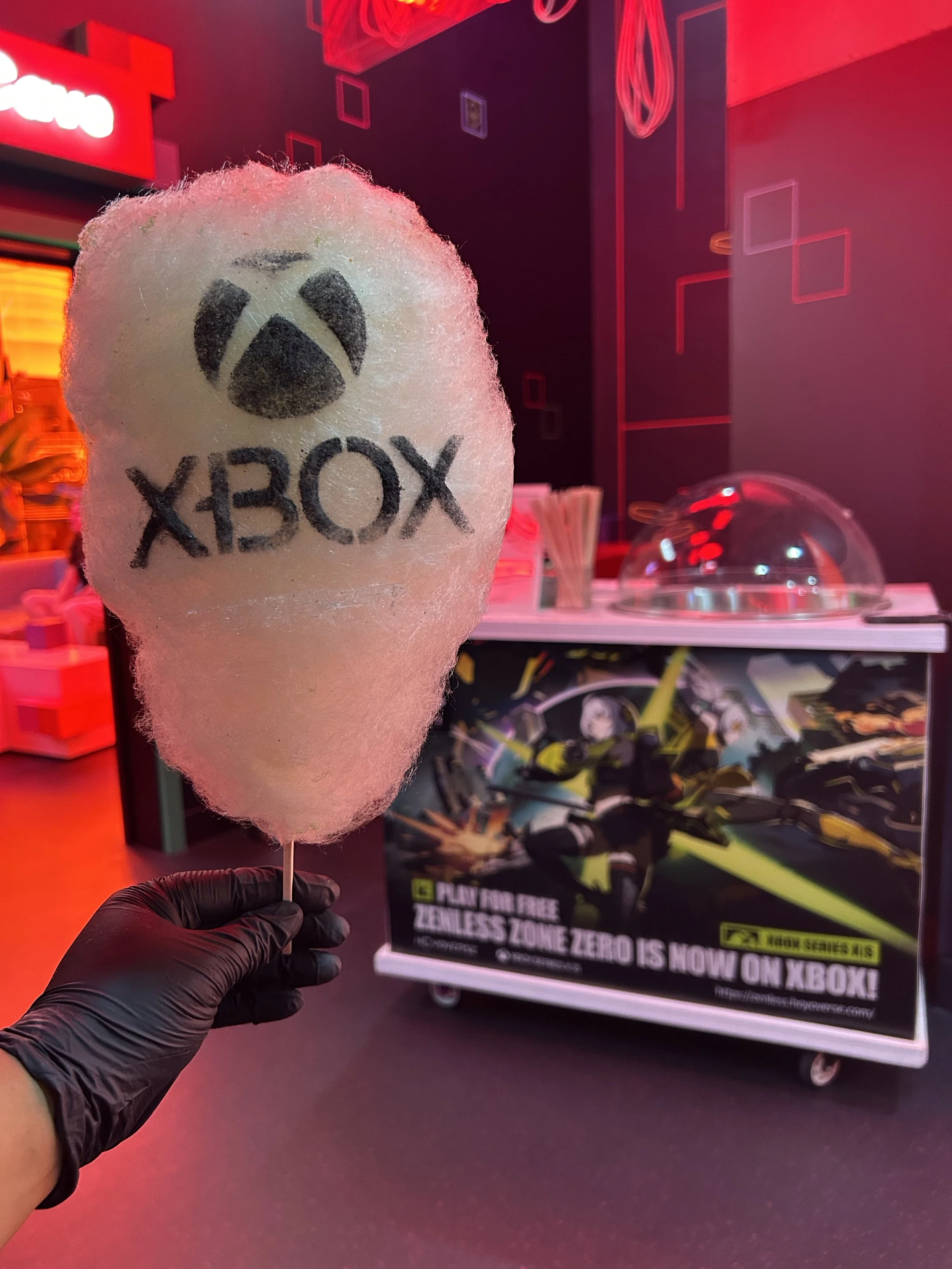 A cotton candy with a logo of a panda and the word 'XBOX' written underneath, being held by a person wearing a black glove, with a gaming store or electronics store in the background featuring Xbox promotional posters and illuminated signage.