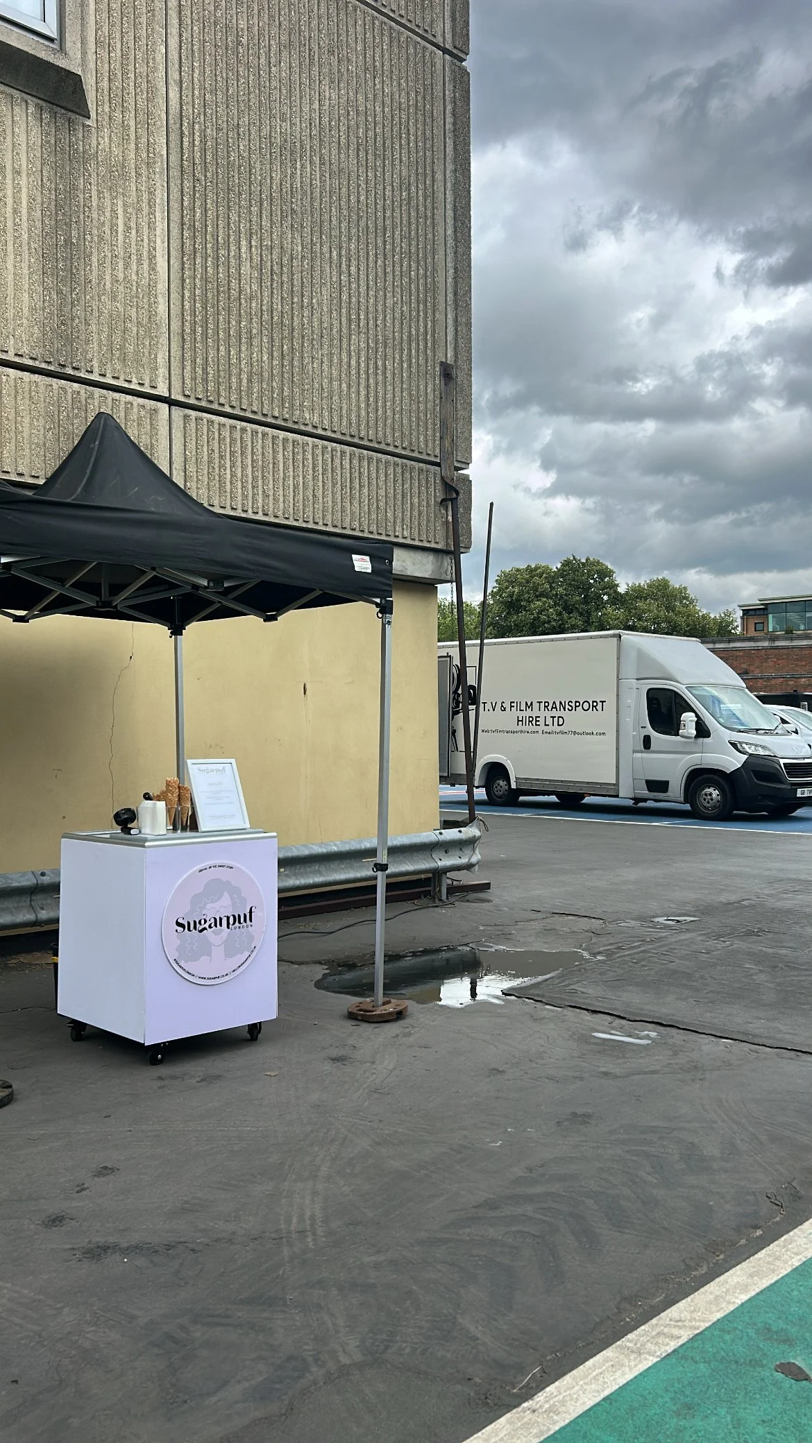 An outdoor parking lot with a small food stand labeled 'Sugarpuff' in front of a black tent. A white delivery van labeled 'T.V & Film Transport Hire Ltd' is parked in the background. The sky is cloudy.