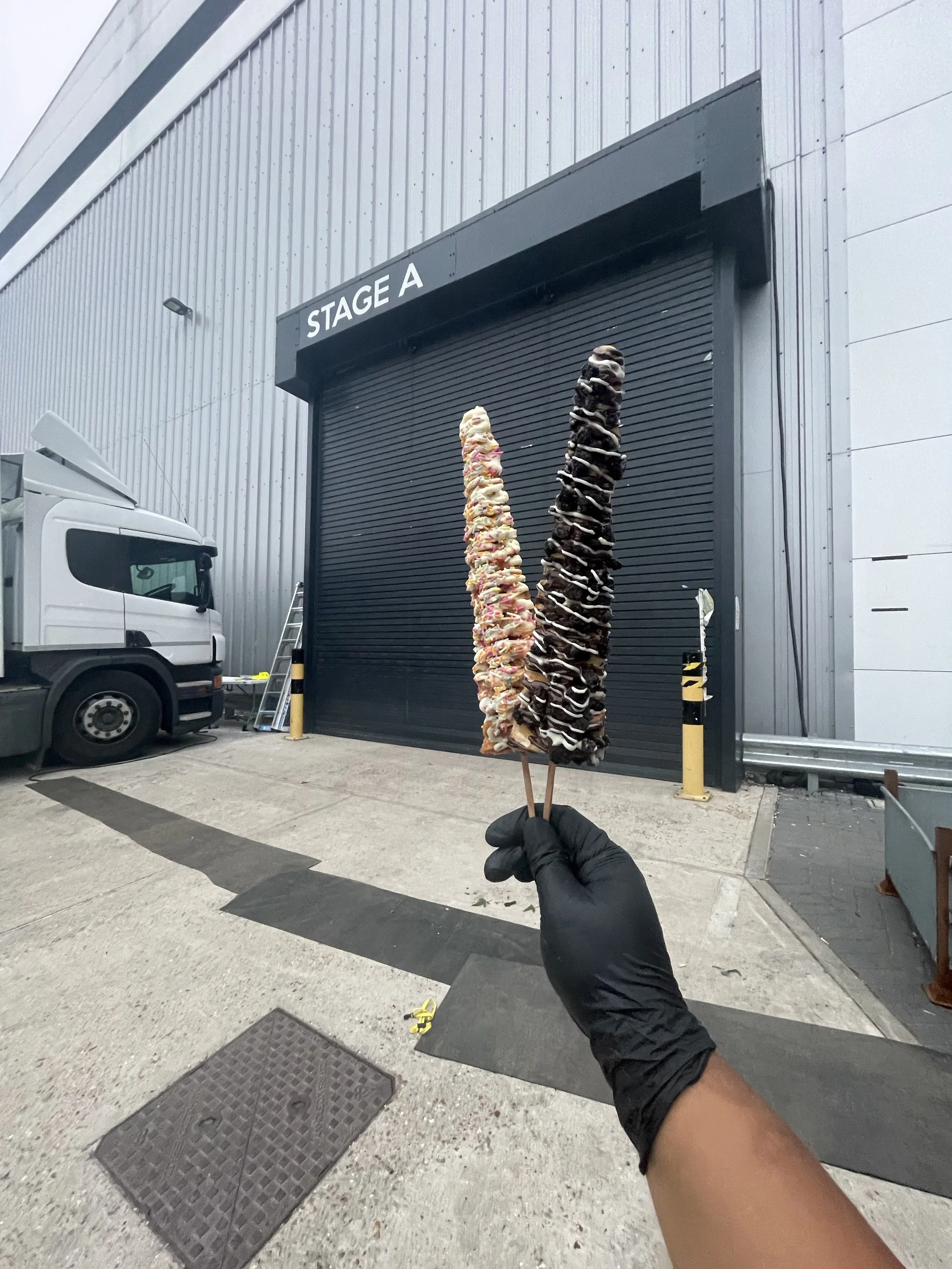 Two large rolled ice cream treats, one with white and pink toppings, and the other with chocolate toppings, are held up outdoors in front of a grey industrial building with a closed black roll-up door labeled "STAGE A." A white truck is parked on the