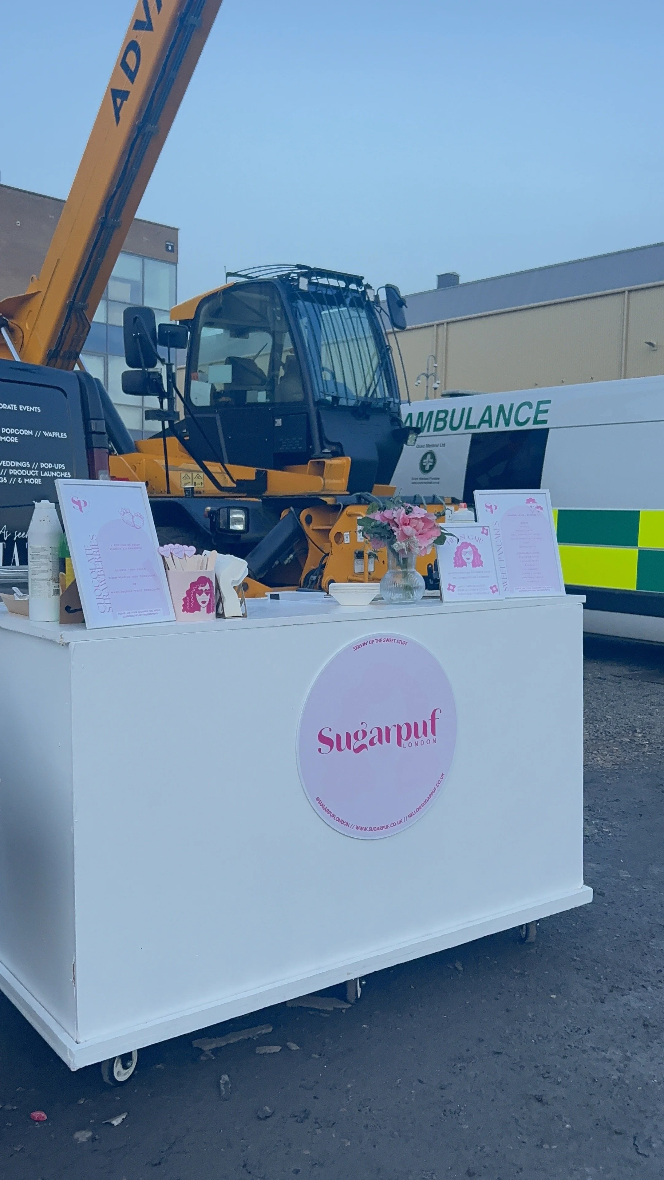 A white food cart with pink signage for Sugarpuff London, featuring couplets of pink and white notes, a small vase of pink flowers, and an attached tentatively transparent dose bottle. Behind it, there is a large yellow crane and an ambulance on a ci