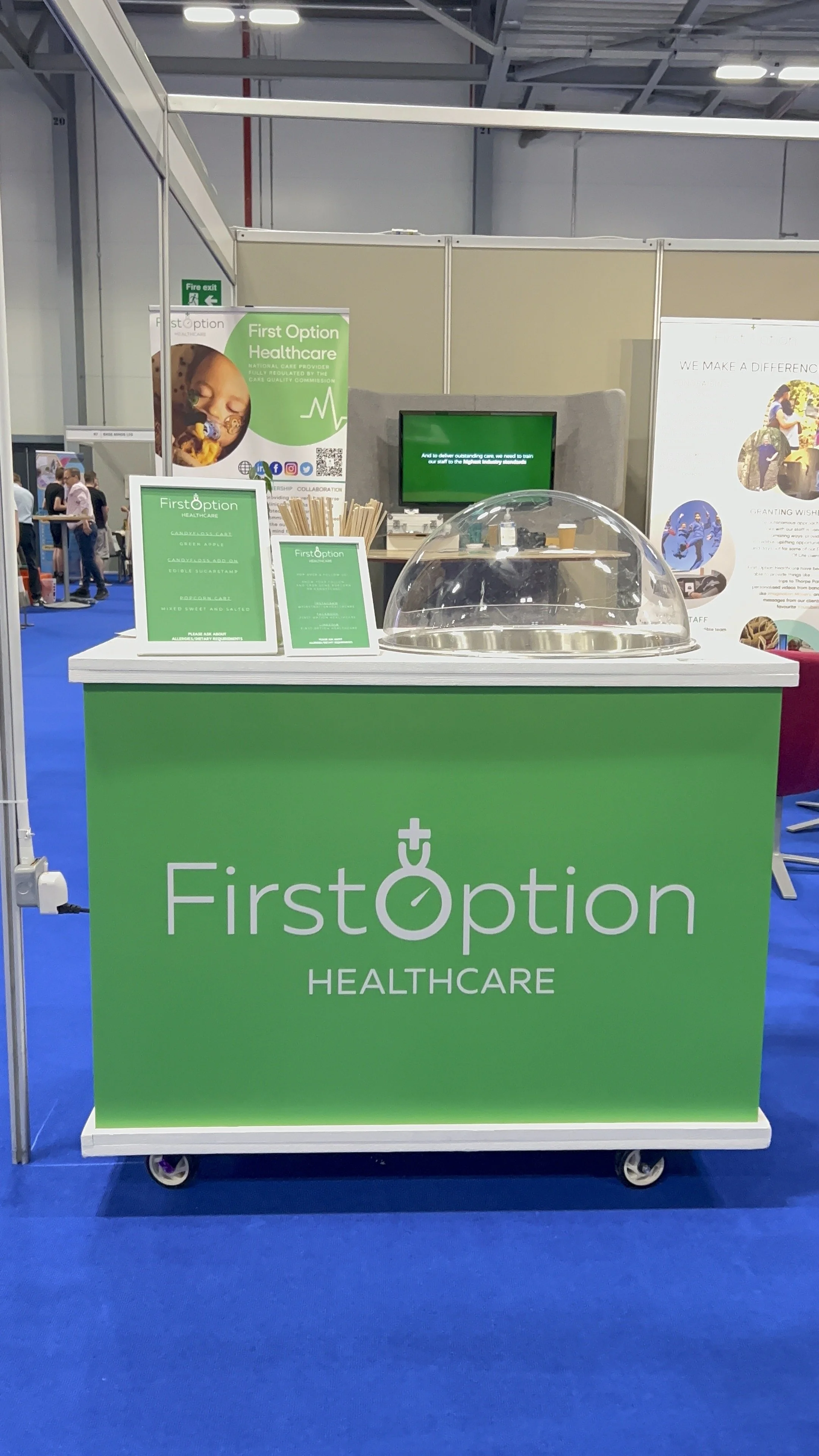 Trade show booth for First Option Healthcare with green branding, a sign displaying product offerings, a computer monitor, and a clear plastic dome on the counter.