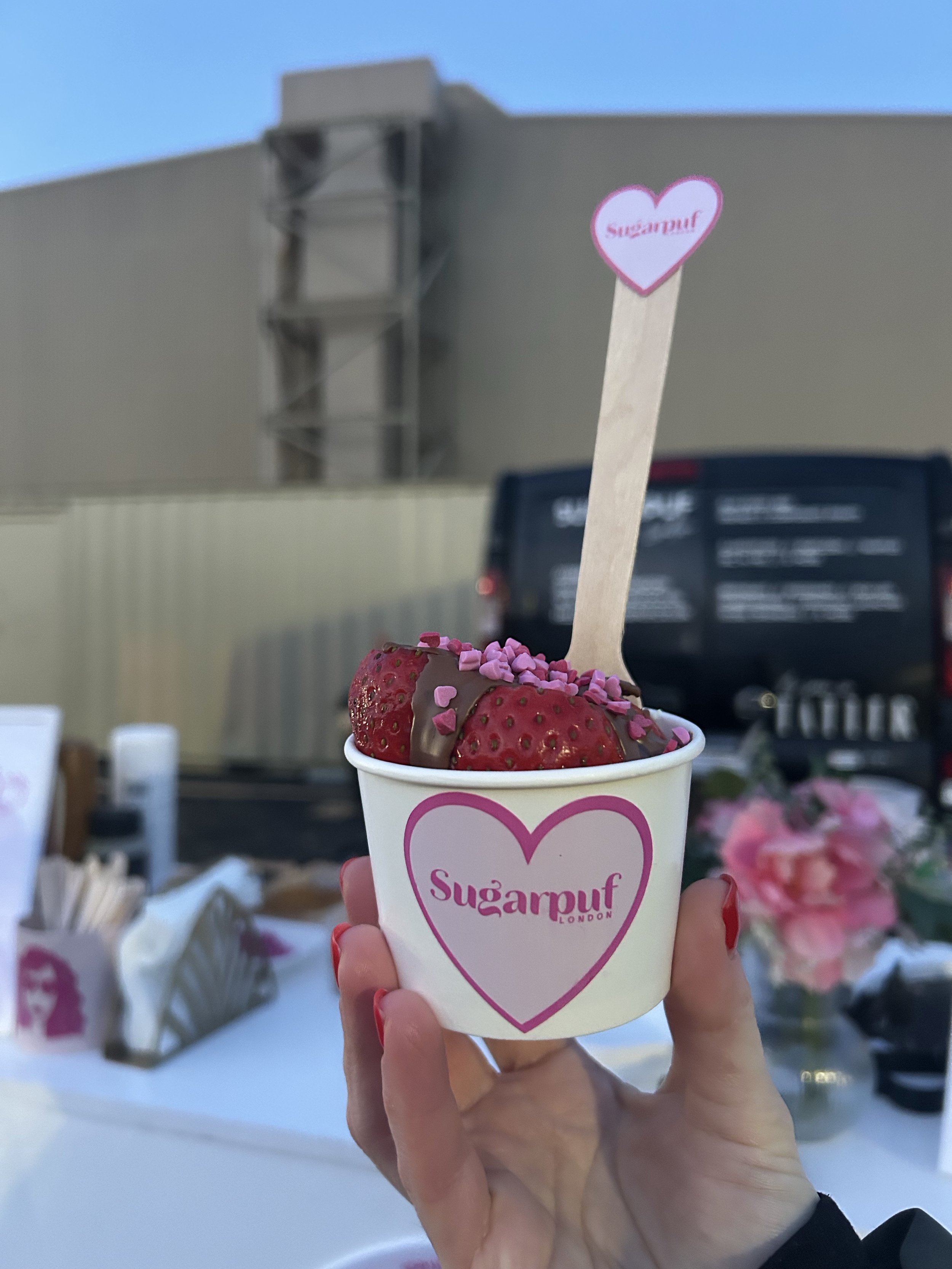 A hand holding a cup of strawberries covered with chocolate and pink heart-shaped sprinkles. The cup has a heart logo with the text 'Sugarpuf London'.