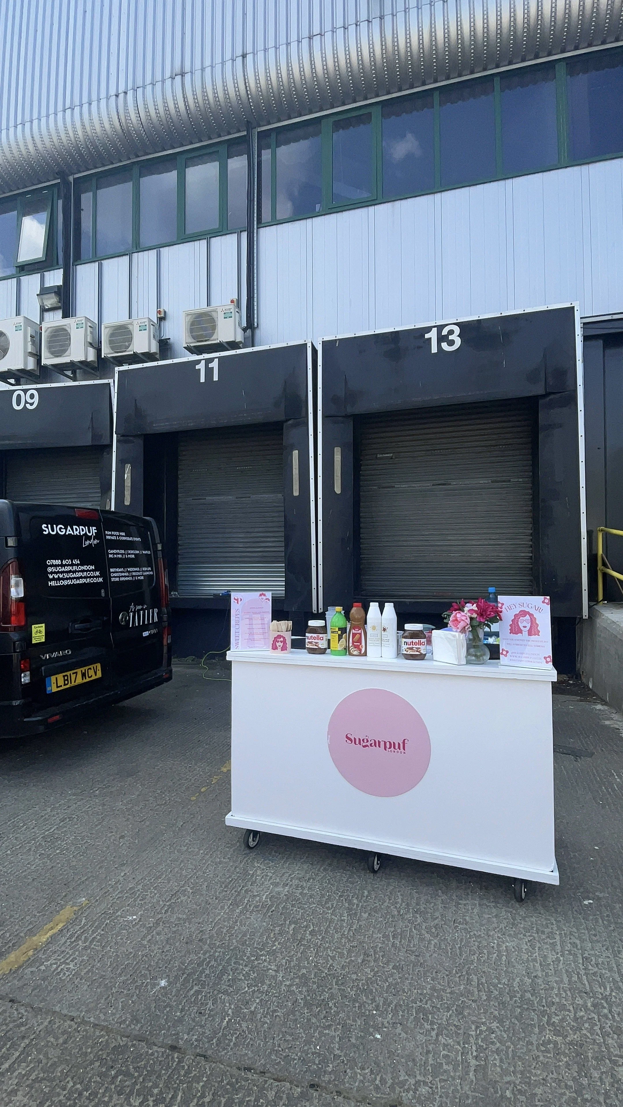 A small mobile stand with the logo 'Sugarpuf' offers various food items including jars and bottles, set up in front of a closed warehouse dock with black doors numbered 11 and 13, in an outdoor parking area.