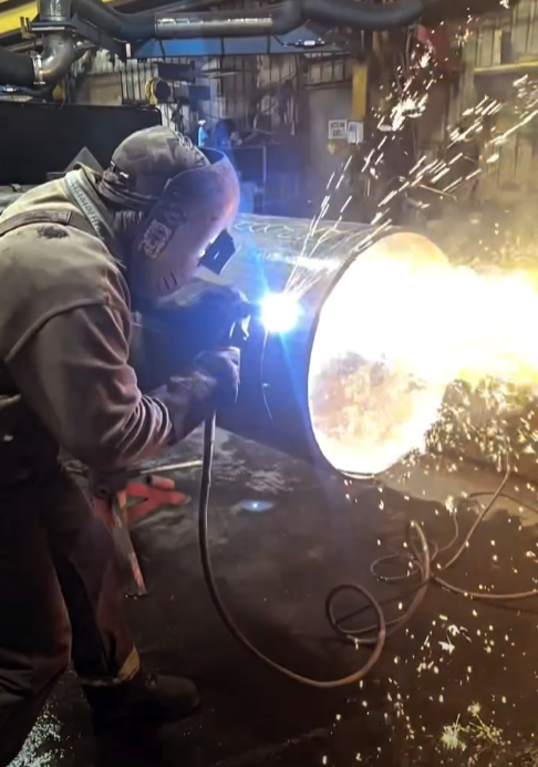 A person welding a metal pipe in an industrial workshop, with sparks and bright light from the welding process.