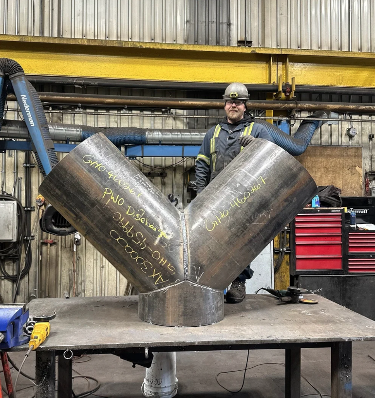 A welder is posing behind a large metal Y-shaped pipe with writing on each side, in an industrial workshop with tools and equipment in the background.