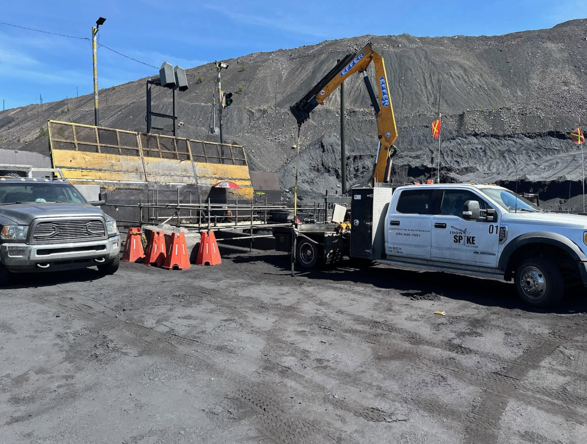 Construction site with a white service truck equipped with a crane, a gray pickup truck, orange safety cones, and a dirt hill in the background under a blue sky.
