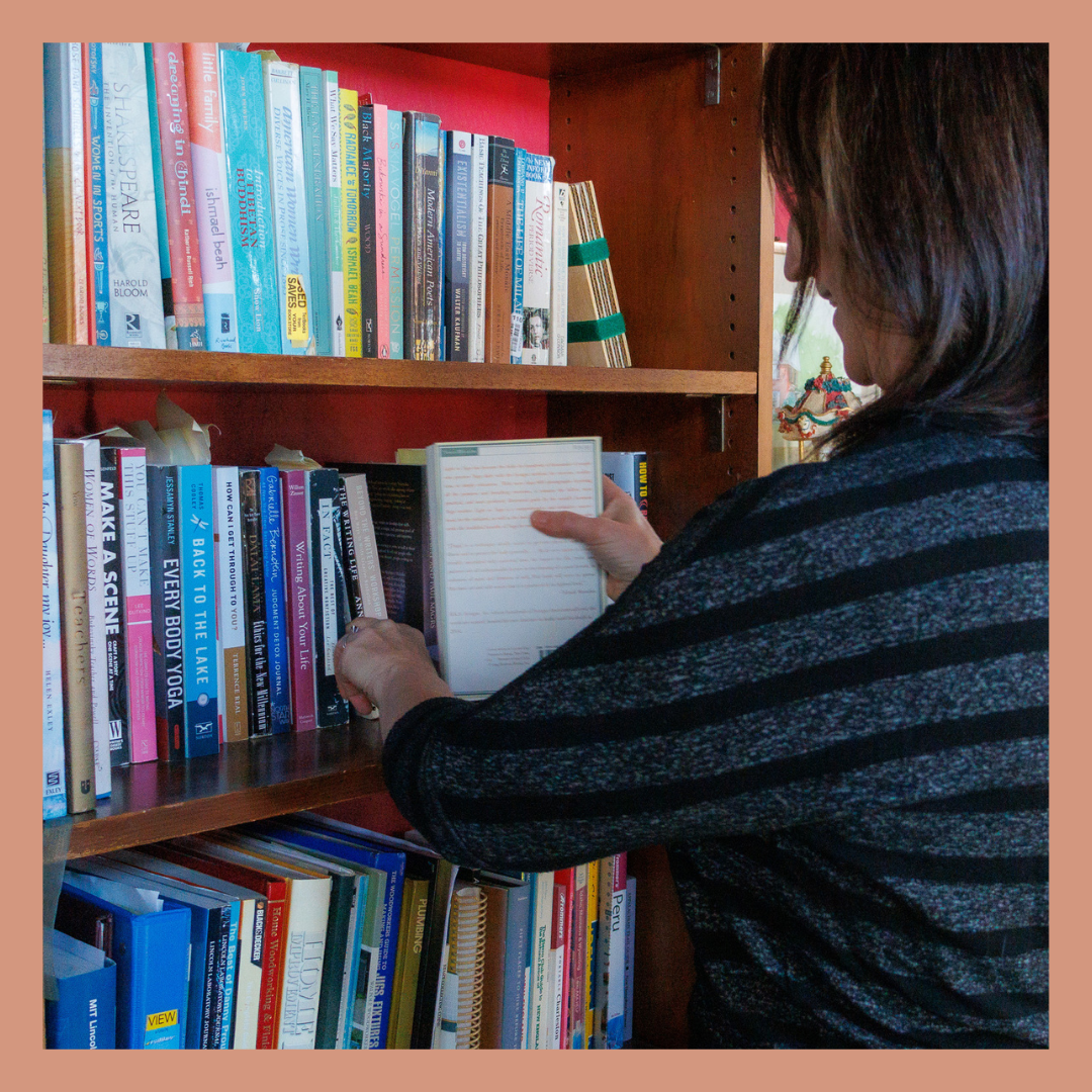 A woman with dark hair and glasses browsing books on a wooden bookshelf, holding an open book.
