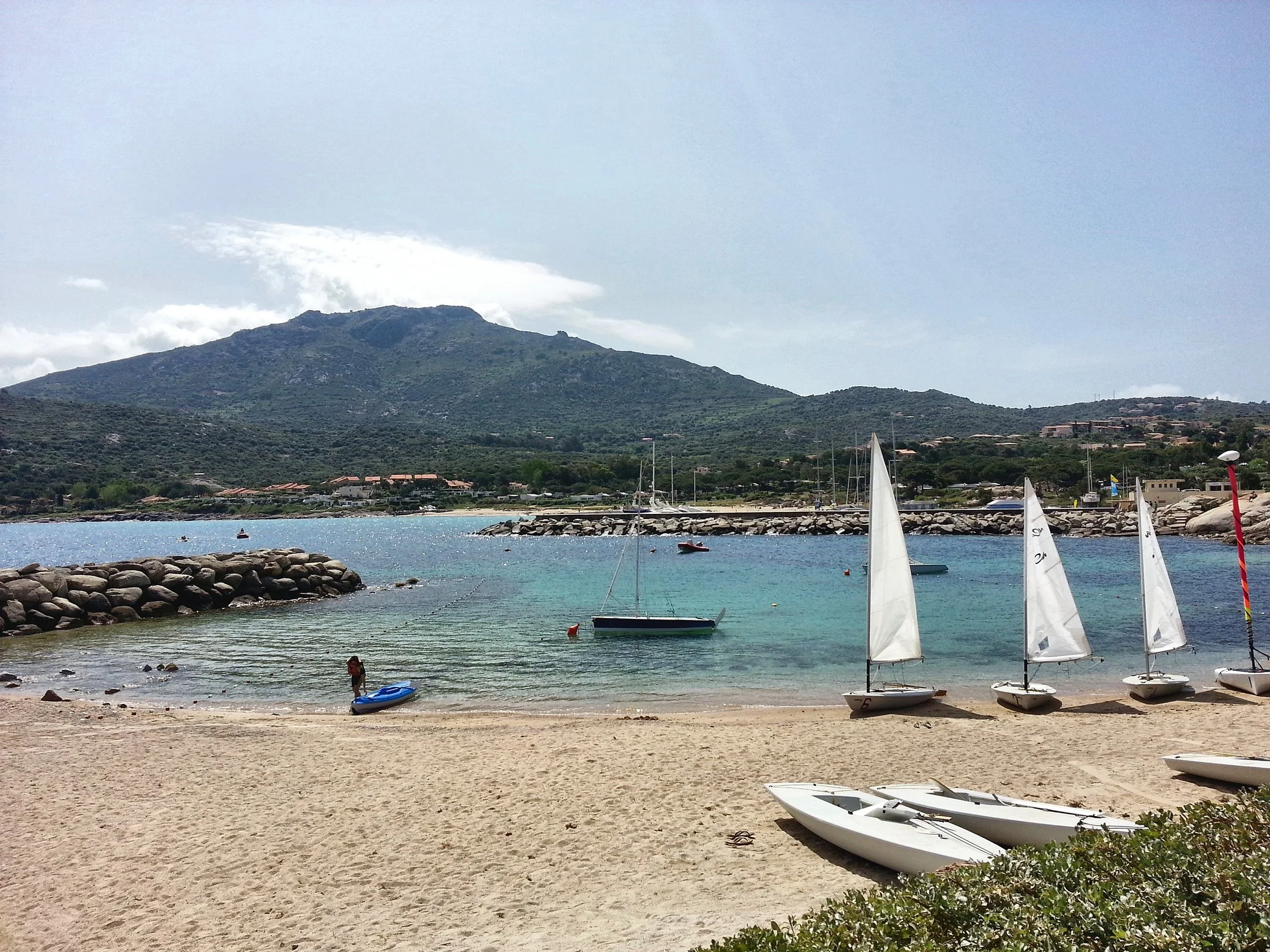 The Club Med Sant’Ambroggio beach in Corsica, with sailing dinghies on the sand, a breakwater and the hills behind