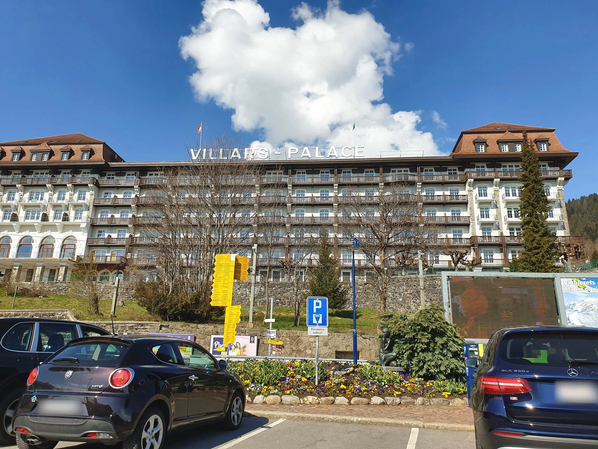 The long white facade of the Villars Palace, with its rooftop sign and wooden balconies, above a car park and spring flower beds under a blue sky