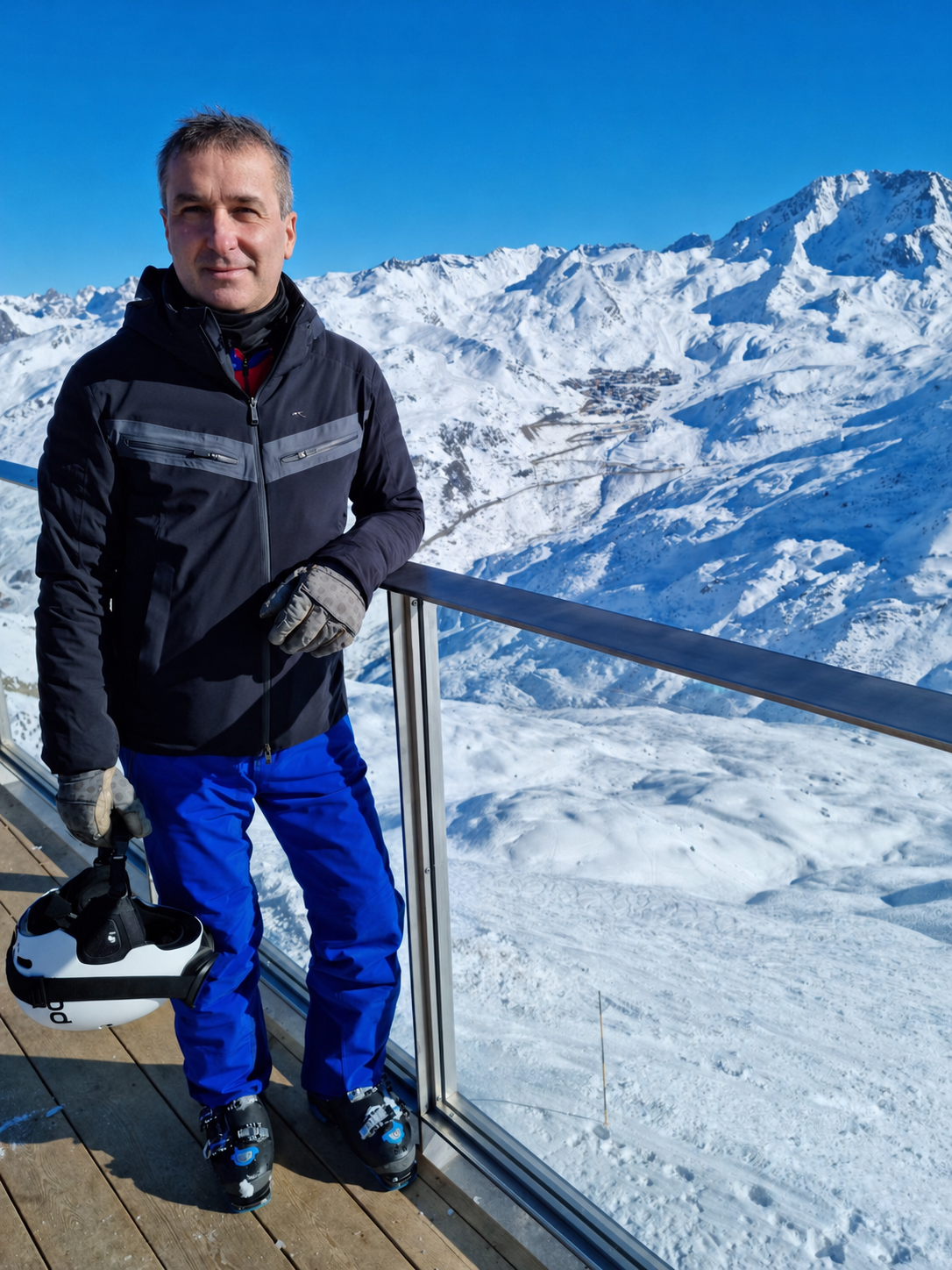 Man dressed in ski gear standing on a snowy mountain deck with a helmet in hand, overlooking a snow-covered mountain range with ski slopes and a small village in the distance under a clear blue sky.