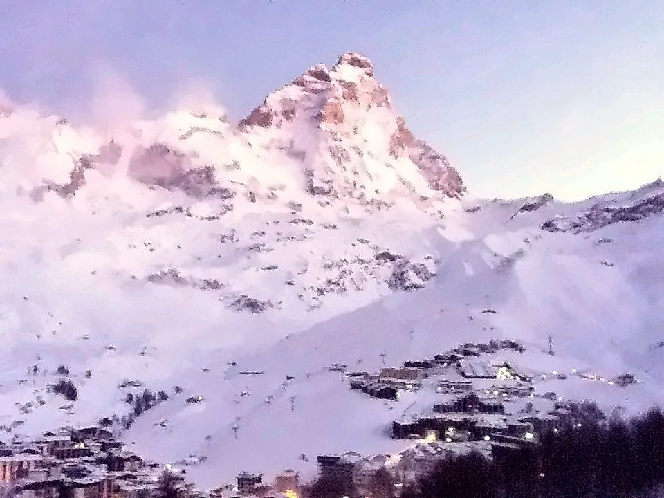 The Matterhorn lit pink at dawn above snow-covered slopes, with the lights of Breuil-Cervinia below