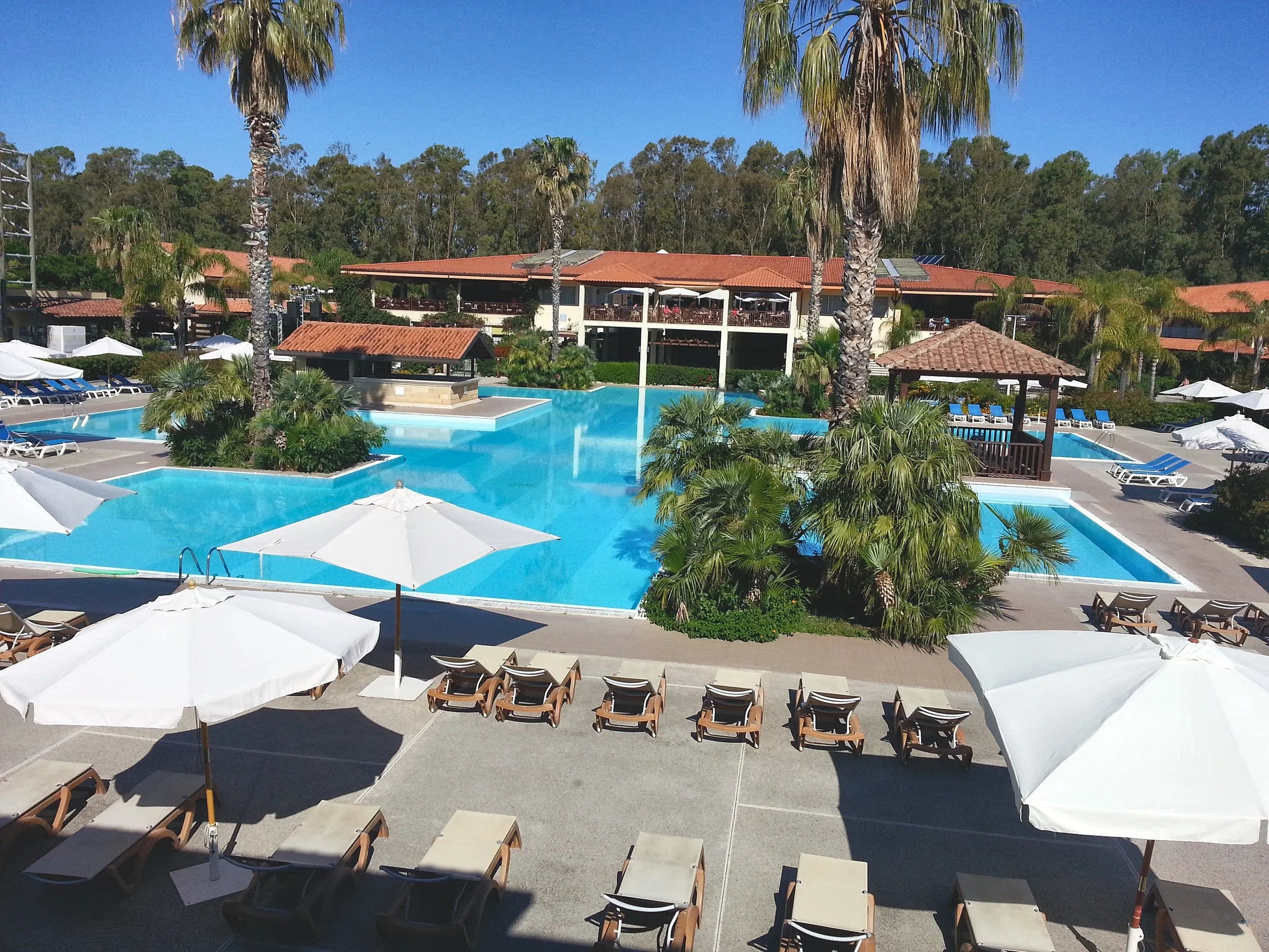 La piscine principale du Club Med Napitia, avec palmiers, transats, parasols blancs et le bâtiment principal au toit rouge devant la pinède