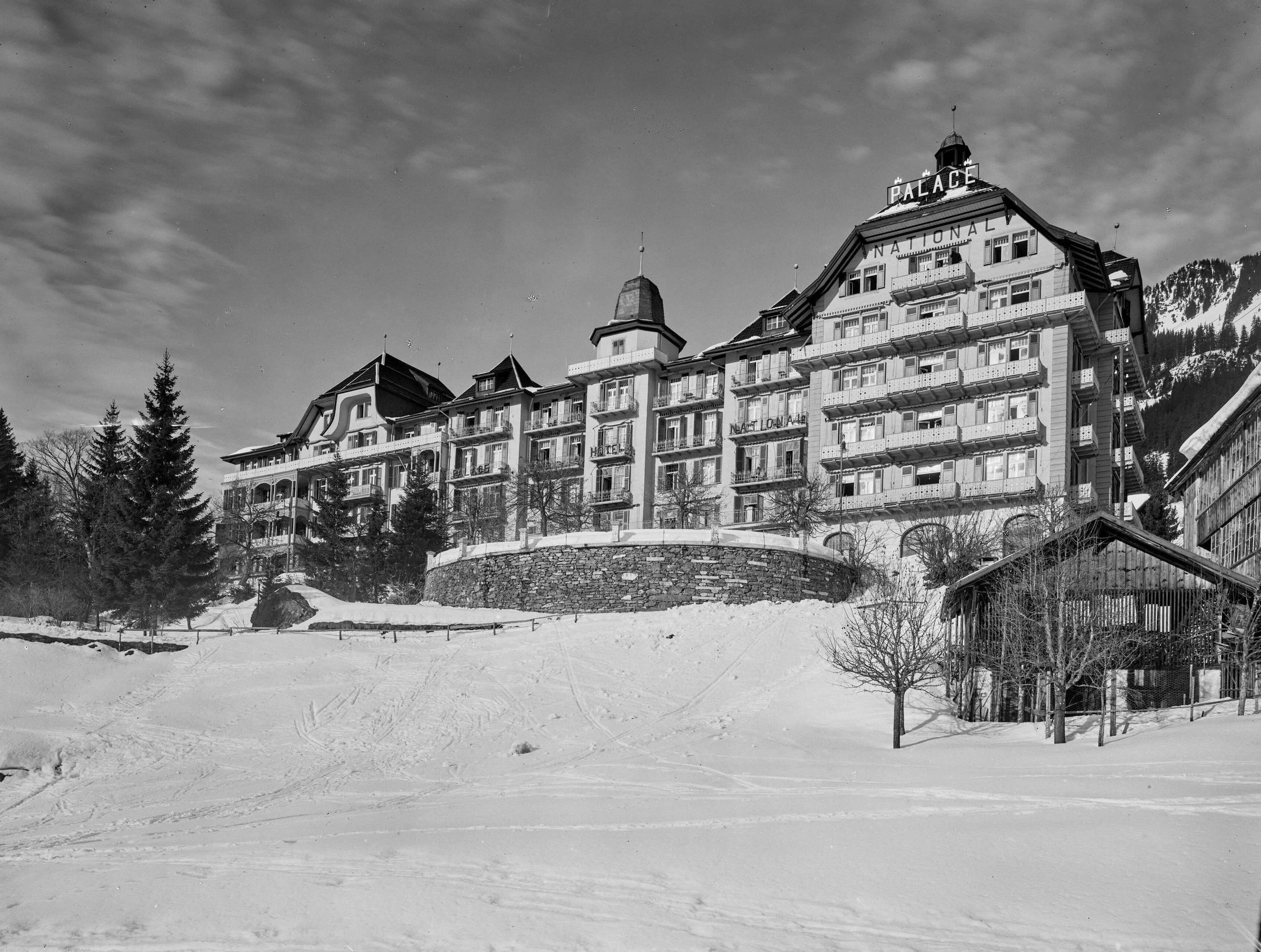 Photographie en noir et blanc du Palace de Wengen en hiver 1923 : un long bâtiment à nombreux balcons, avec une tour à dôme et une haute aile portant l’inscription Palace National, au-dessus d’une pente enneigée