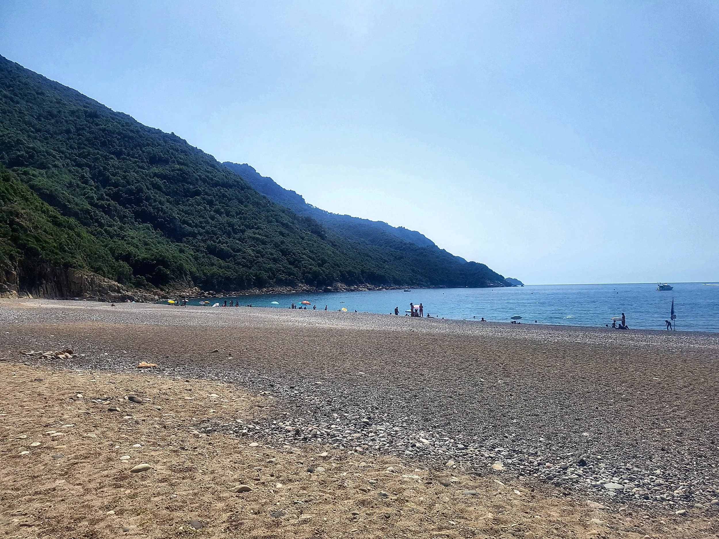 Une large plage de sable et de galets sur la côte ouest de la Corse, avec des collines boisées qui plongent dans la mer, et des baigneurs et des parasols au bord de l’eau