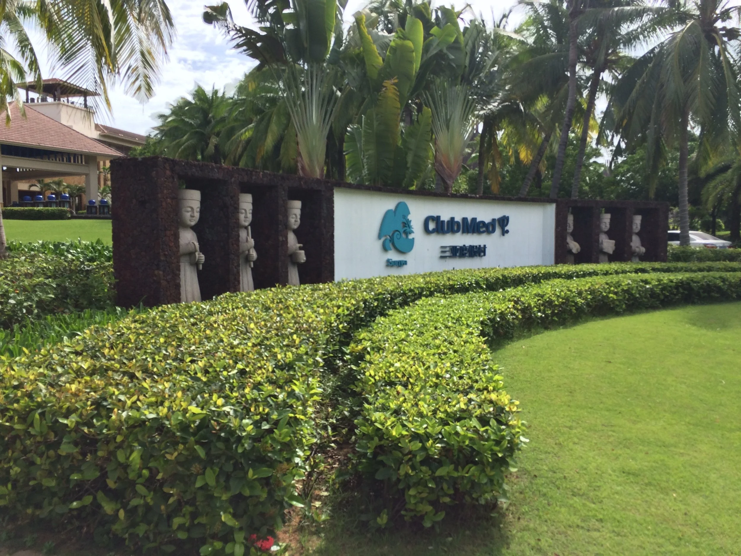 L’enseigne d’entrée du Club Med Sanya, encadrée de statues de pierre, derrière des haies arrondies et des palmiers