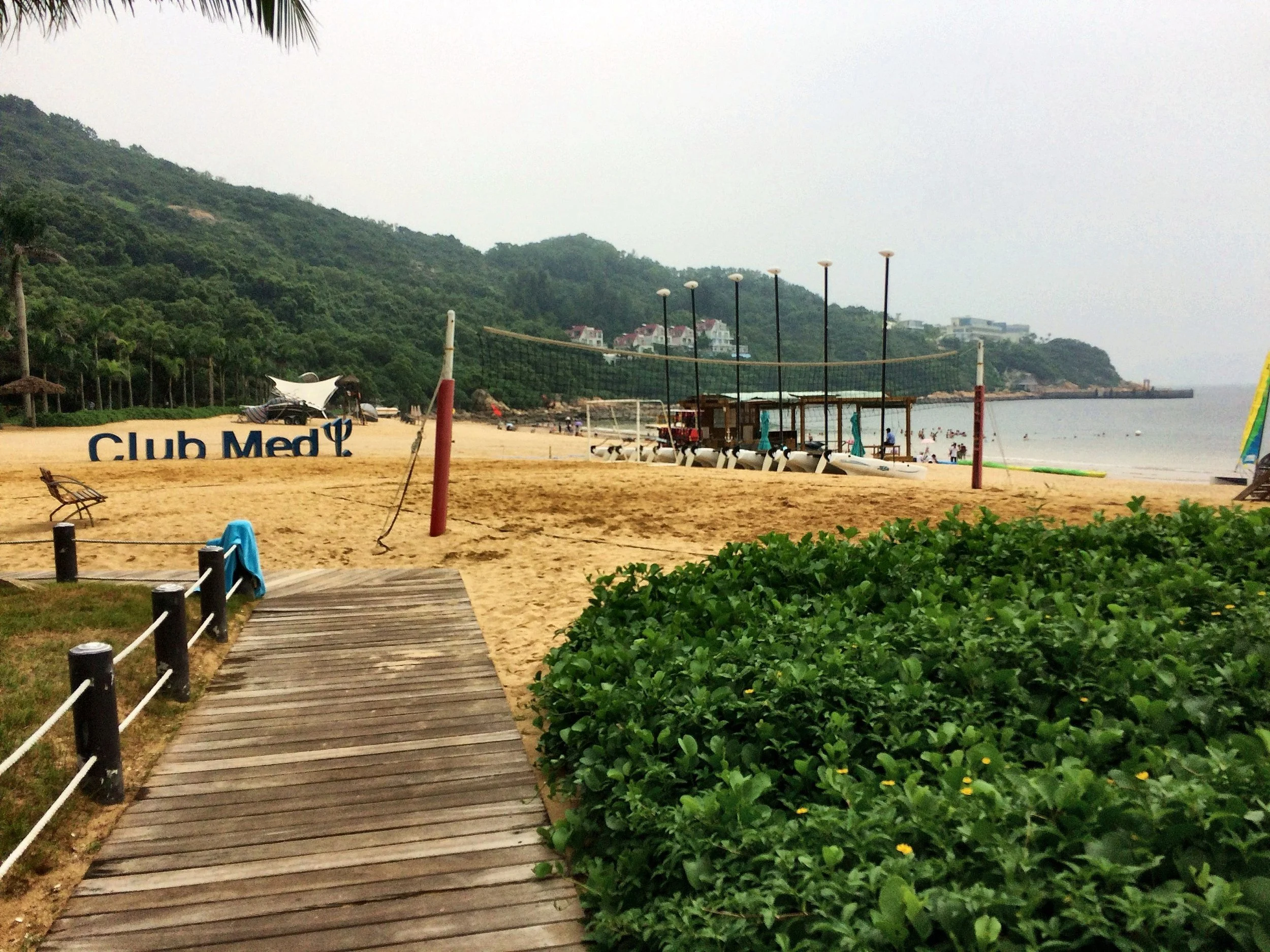 Boardwalk to the beach at the Club Med Dong’ao Island, with the Club Med sign, a volleyball net and catamarans below wooded hills