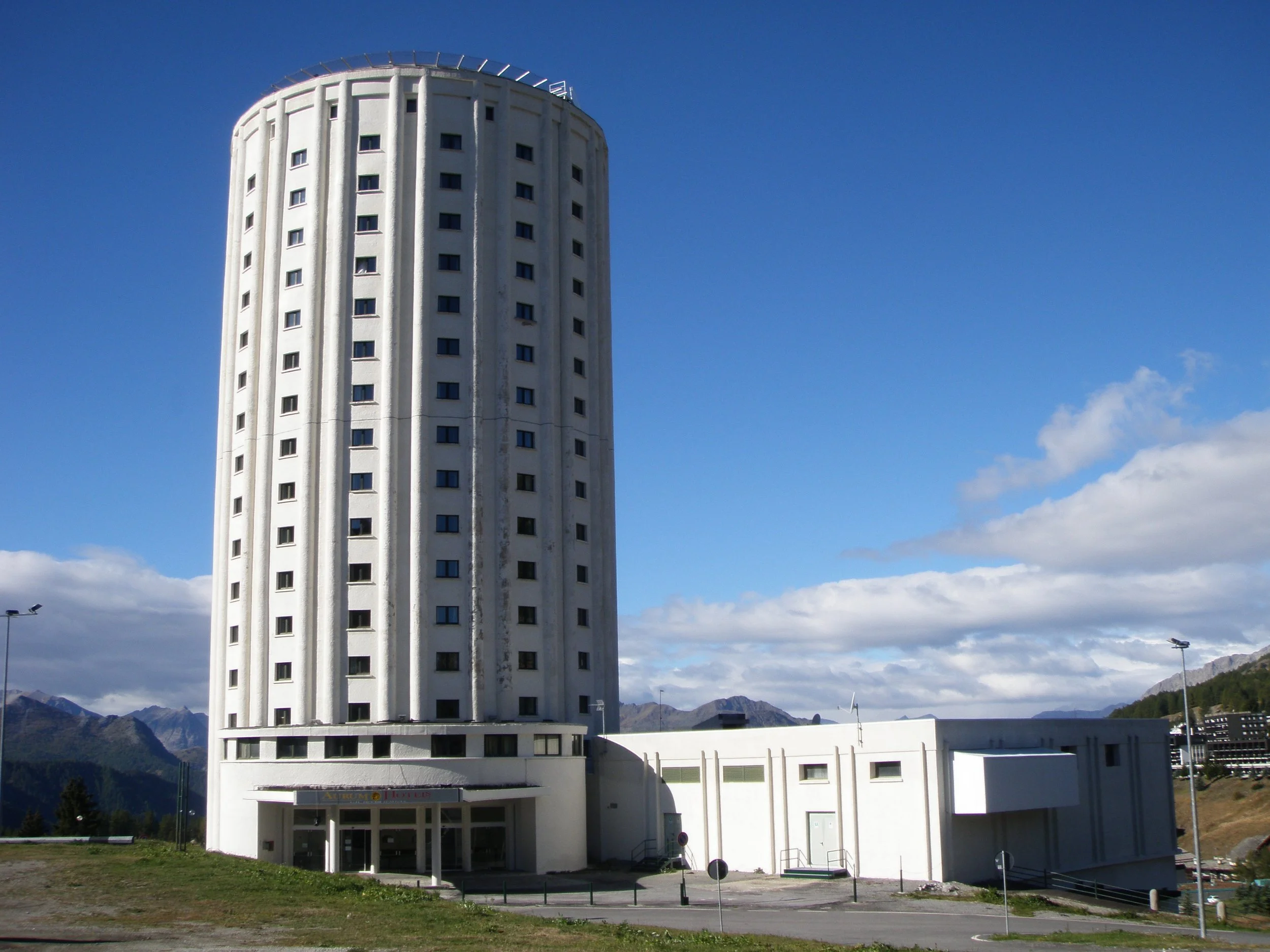 Une haute tour cylindrique blanche, avec un bâtiment bas blanc à côté, sous un ciel bleu à Sestrières