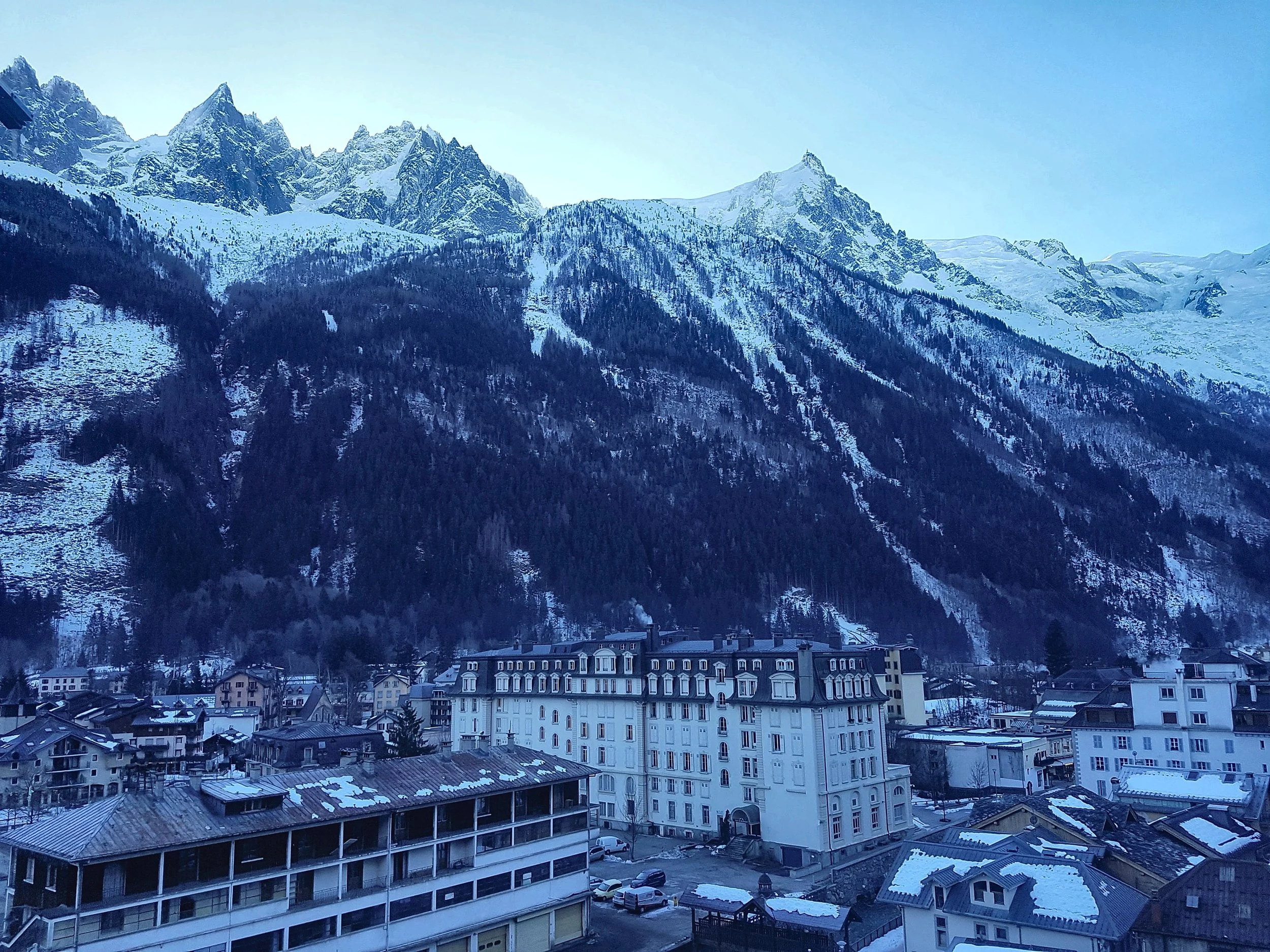 Toits enneigés de Chamonix et grand bâtiment blanc de style palace aux toits mansardés, sous le versant boisé et les aiguilles de granit de Chamonix, dans la lumière bleue et froide de l’hiver