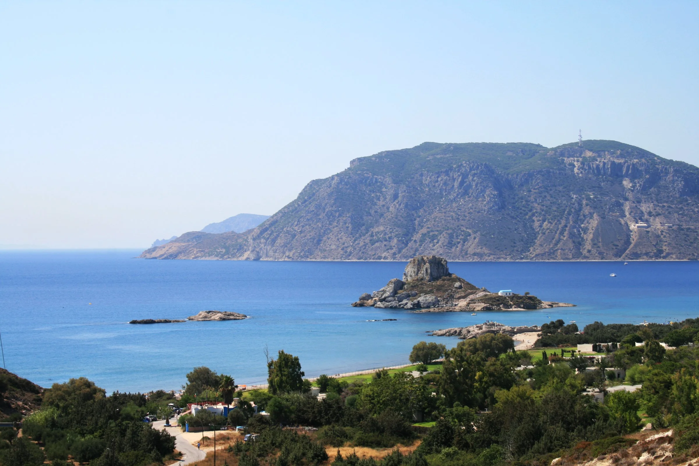Kastri islet, with its small white chapel, off the beach at Agios Stefanos in the Bay of Kefalos, with the mountains of the Kefalos peninsula behind and low buildings among trees in the foreground
