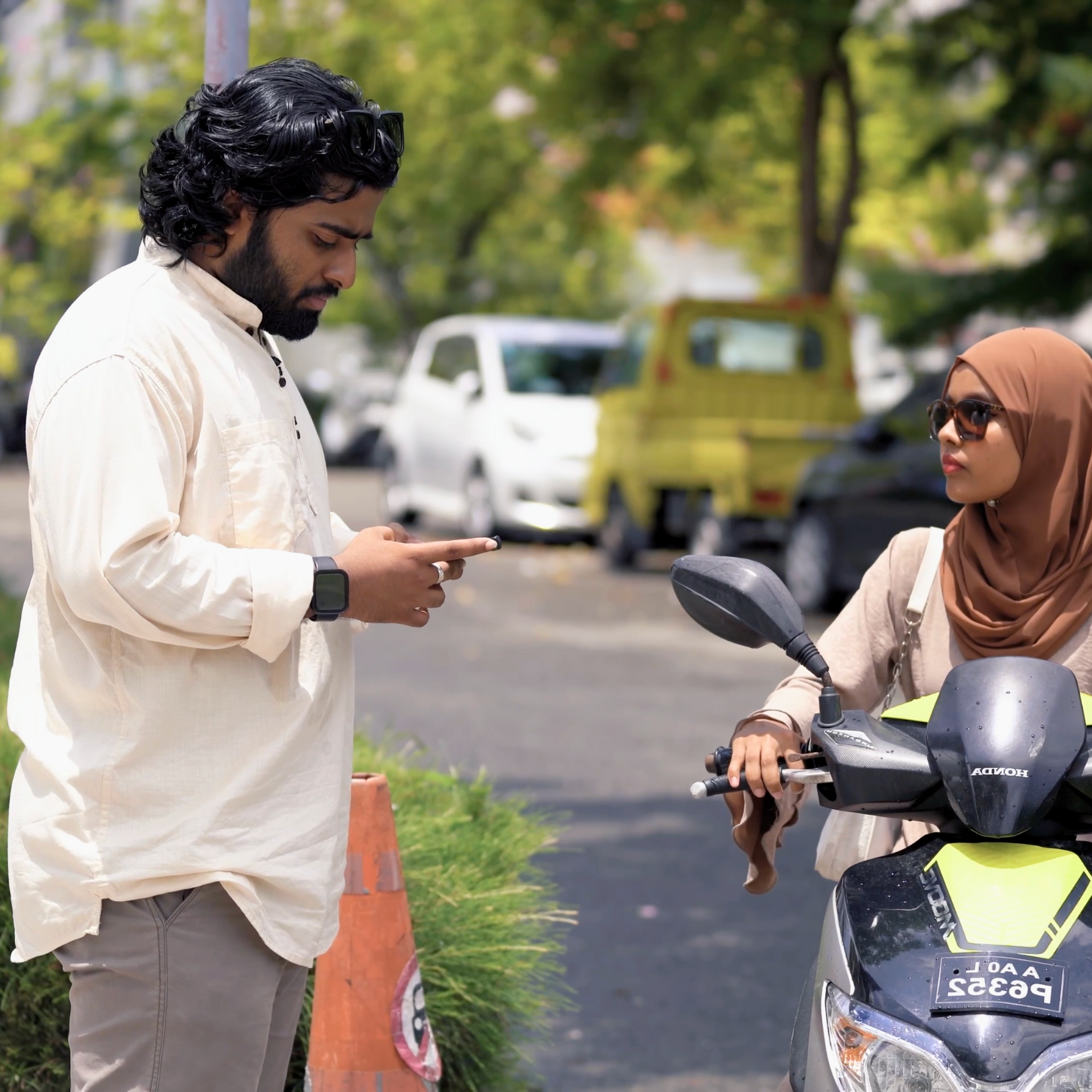 A man with long curly black hair, wearing a beige shirt and a smartwatch, is looking at his phone while standing on the sidewalk. A woman wearing sunglasses and a brown headscarf is sitting on a black and yellow Honda scooter and looking at the man. The background shows parked cars and trees.