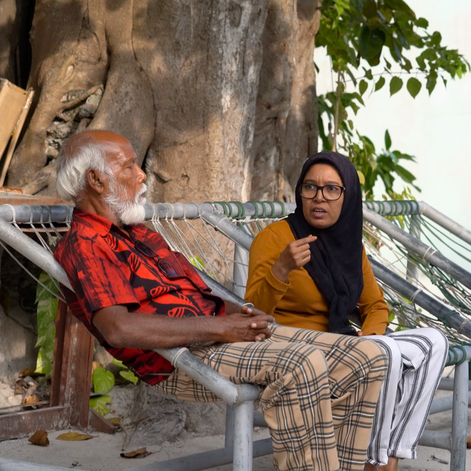 An elderly man with white hair and beard, wearing a red patterned shirt and checkered pants, sitting on a hammock. A woman with glasses and a black hijab, wearing an orange top, is sitting beside him, talking and using hand gestures. They are outdoors with large tree roots and green foliage in the background.