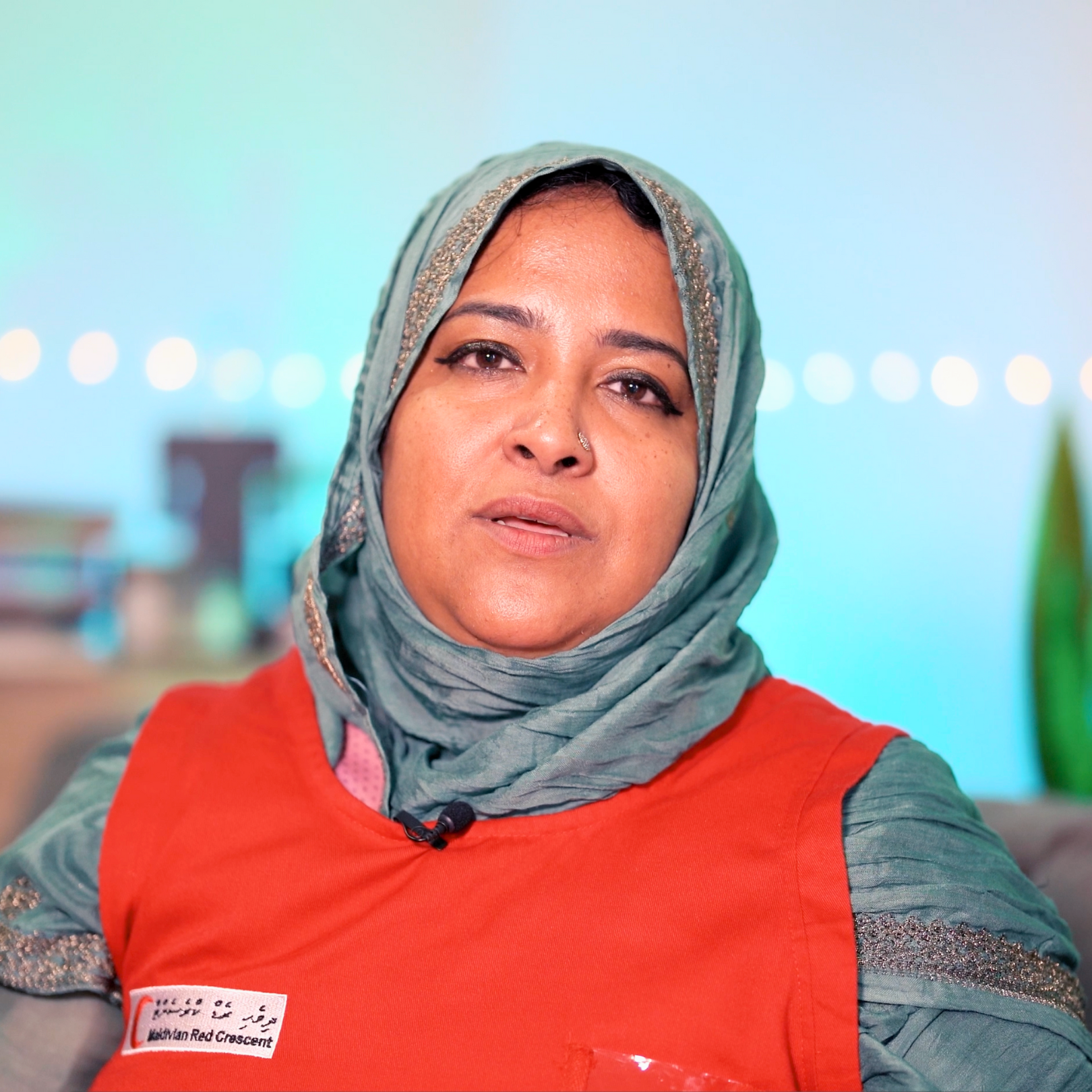 A woman wearing a green headscarf and red vest speaks, with blurred lights and plants in the background.