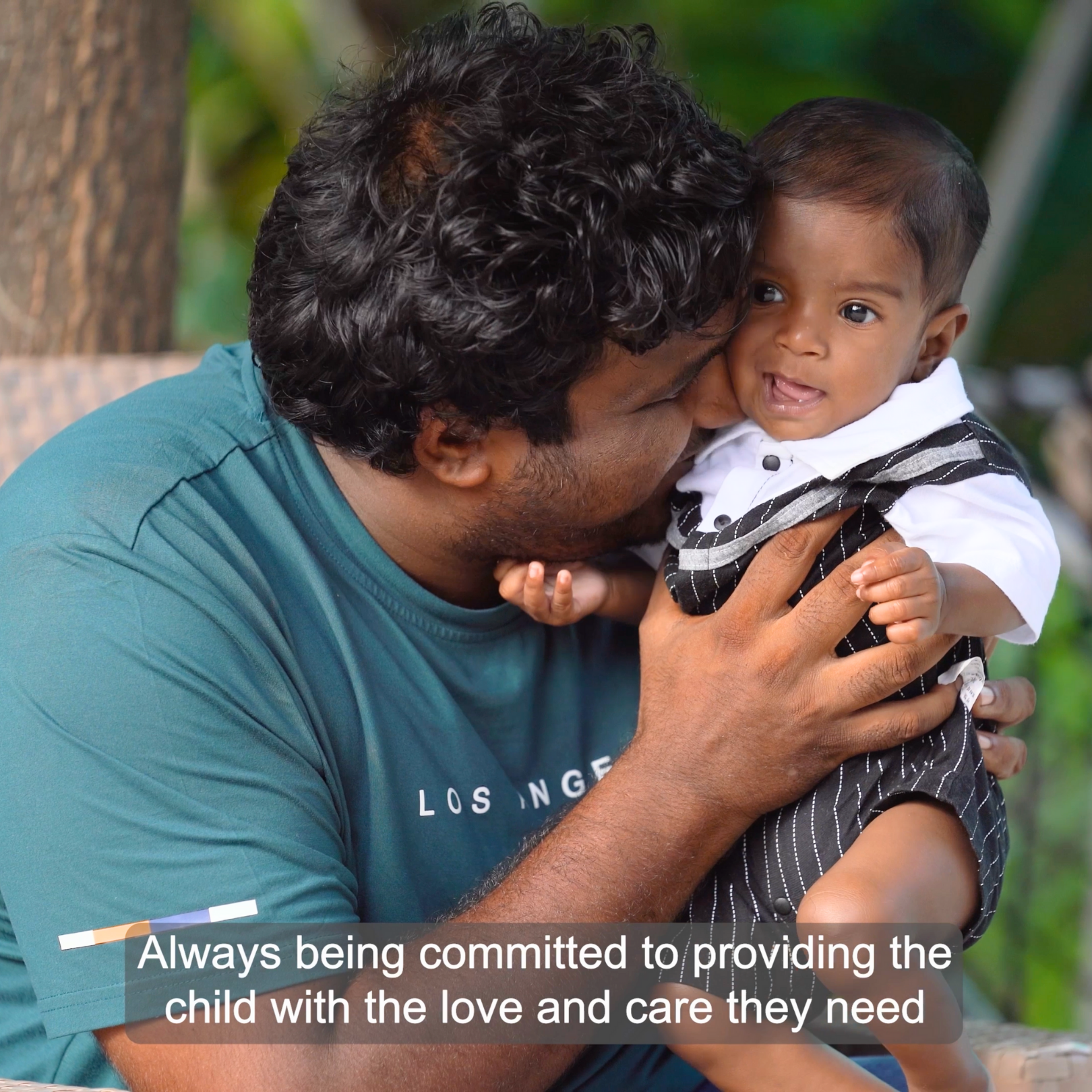 A man cuddling a young boy outdoors, with greenery in the background, showing affection and care.
