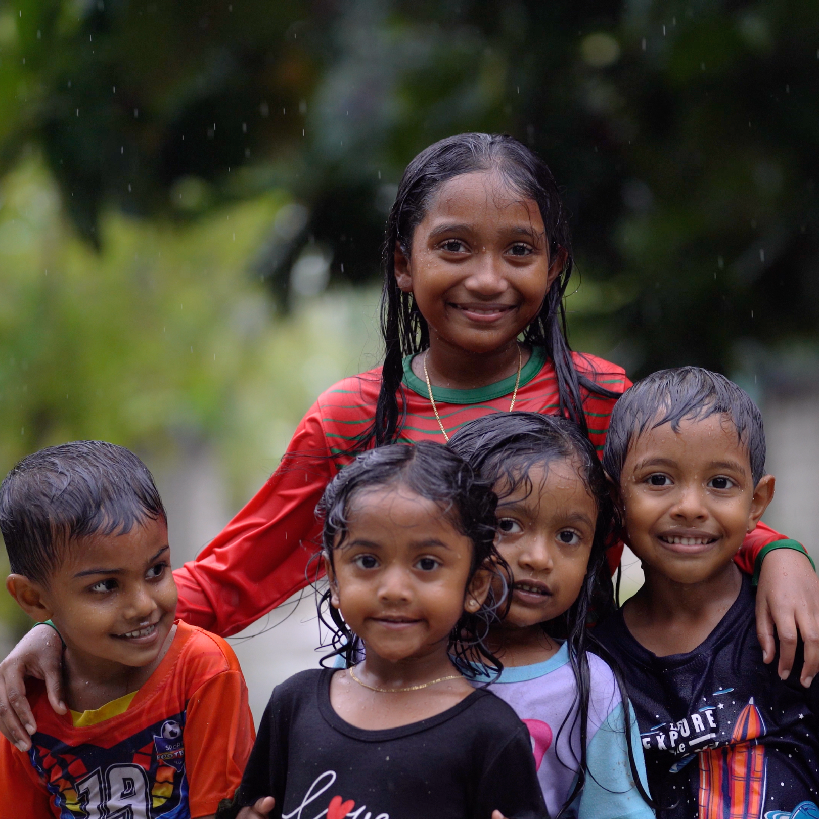 Group of five children playing in the rain, smiling and wet, outdoors with greenery in the background.