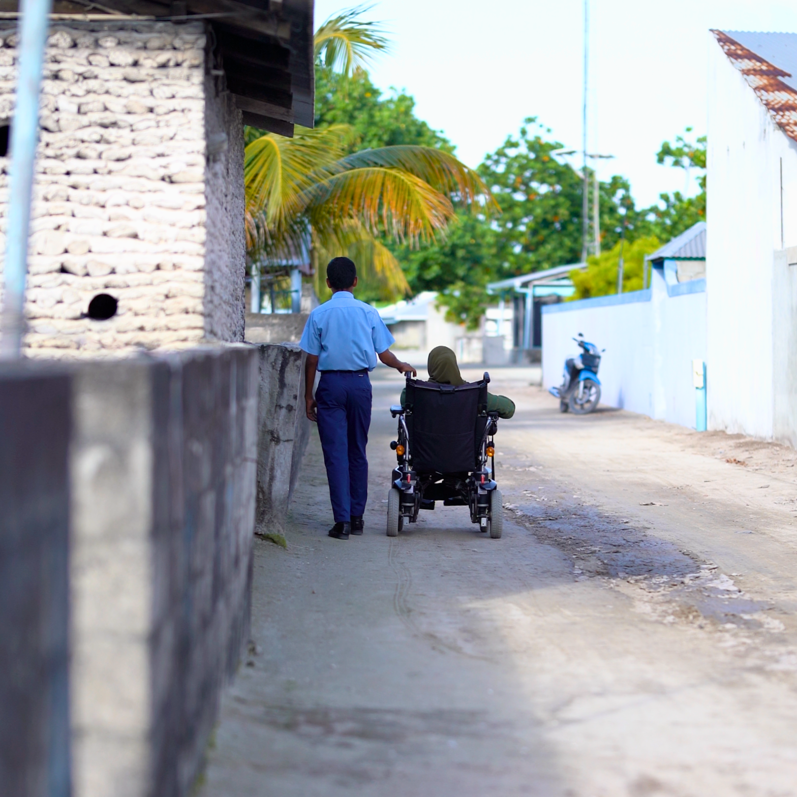 A young man in a blue uniform pushing a woman in a wheelchair along a dirt street in a small neighborhood with houses, palm trees, and a motorcycle parked on the side.