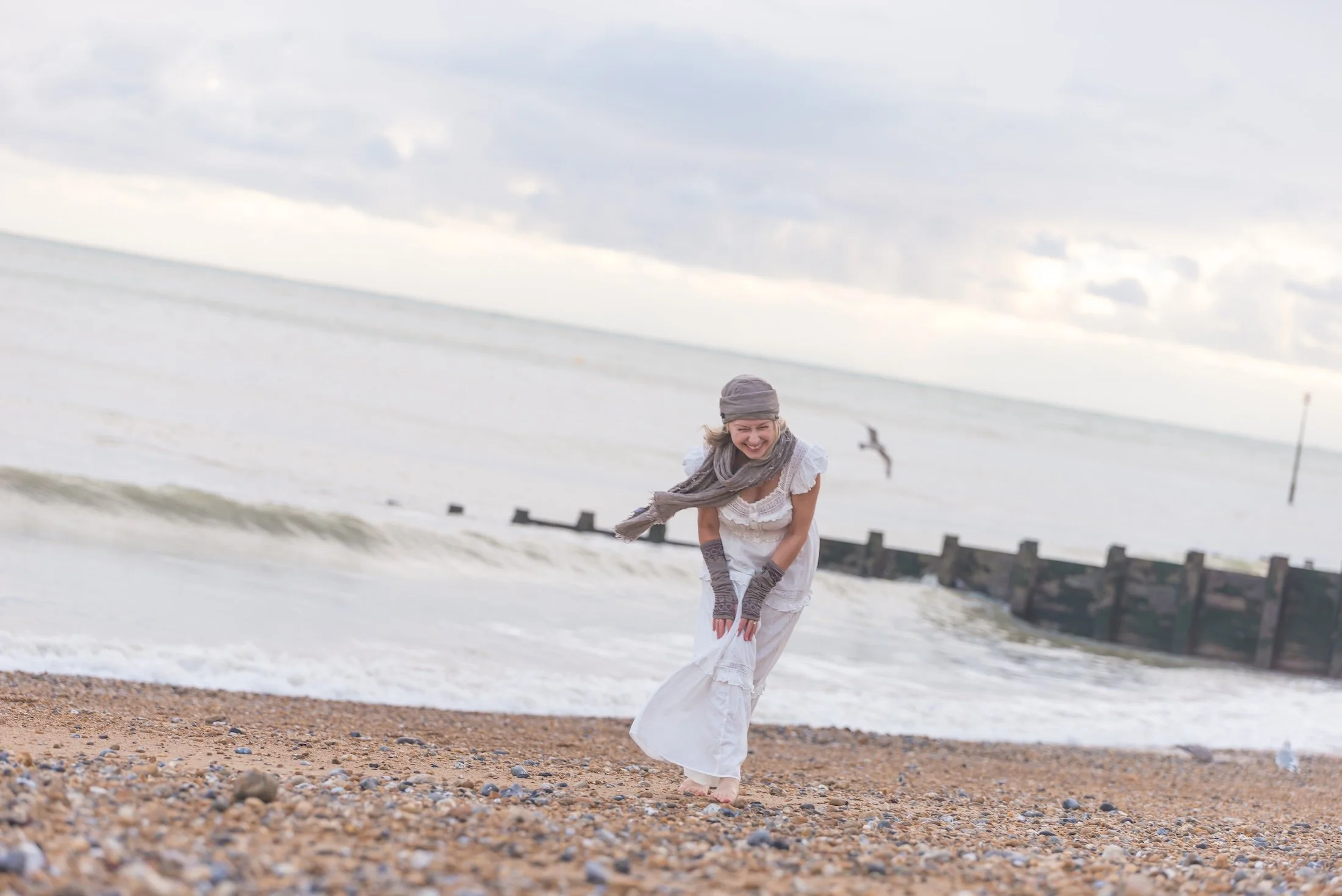 A woman dressed in white with a grey scarf and gloves standing on a pebbled beach, laughing and holding her dress, with waves and a cloudy sky in the background.