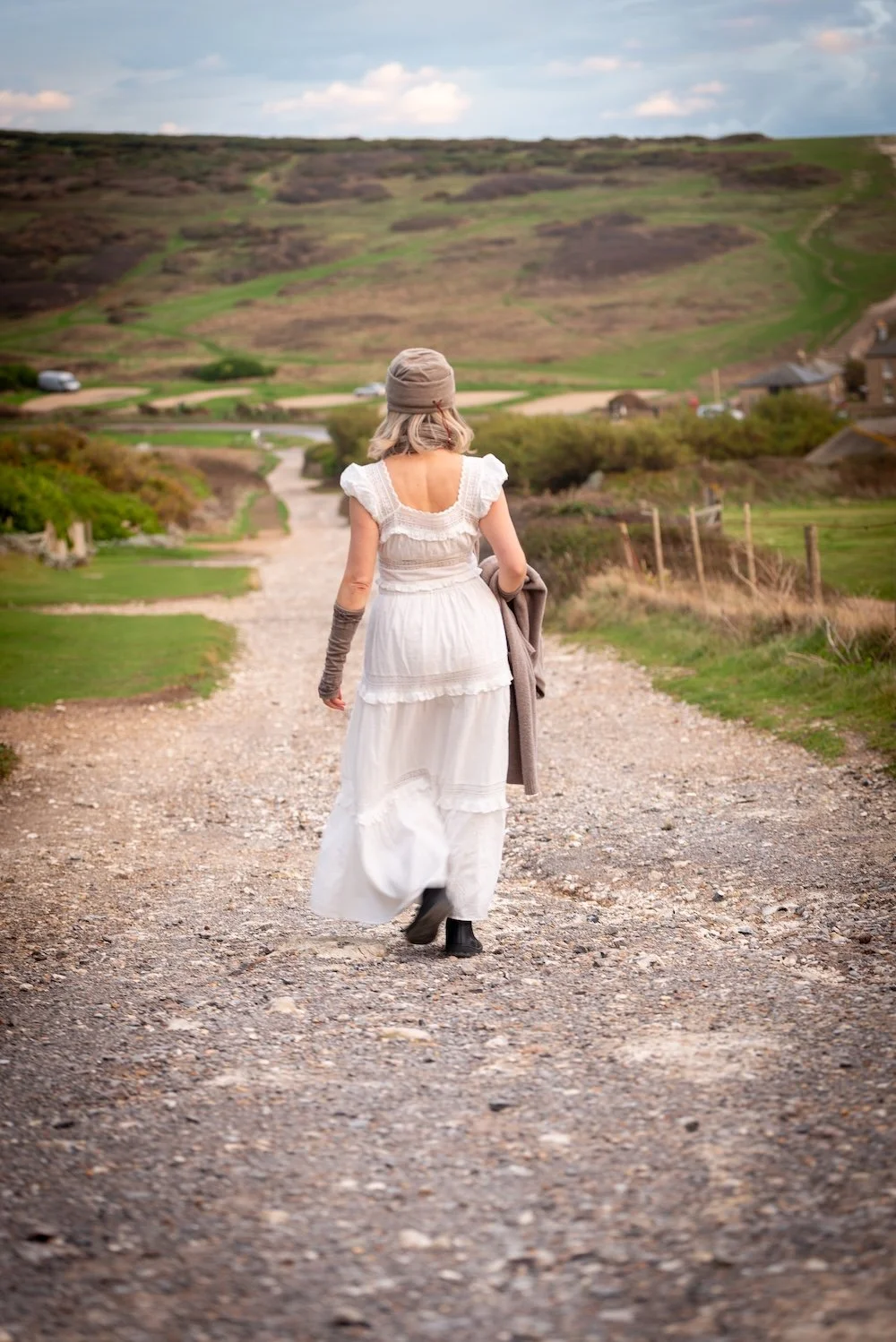 Woman in a white dress walking down a gravel road in a rural landscape with green fields, fences, and hills in the distance.