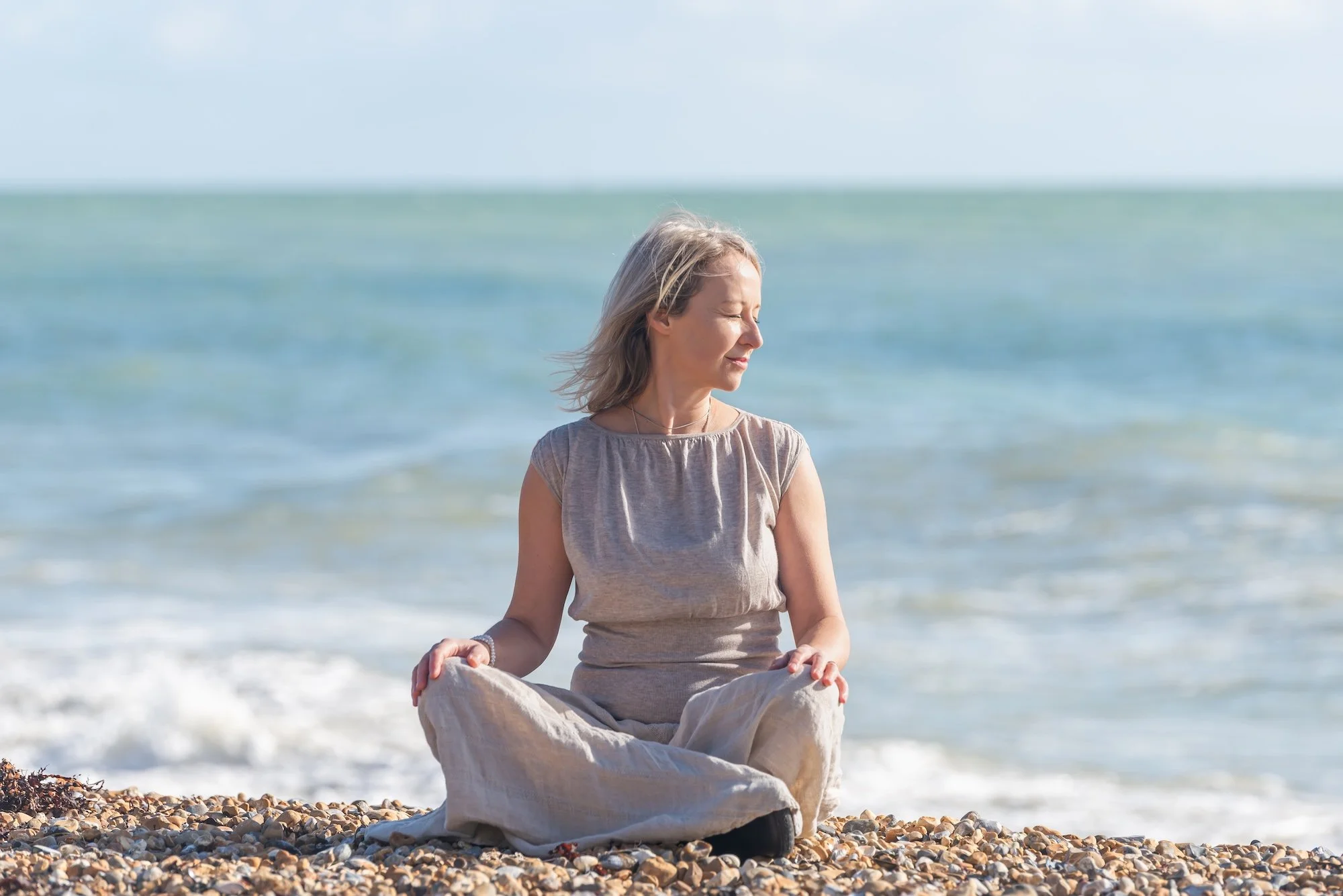 Woman meditating cross-legged on a pebbled beach, facing the sea, eyes closed, sunlight on her face; embracing bodymind harmony.