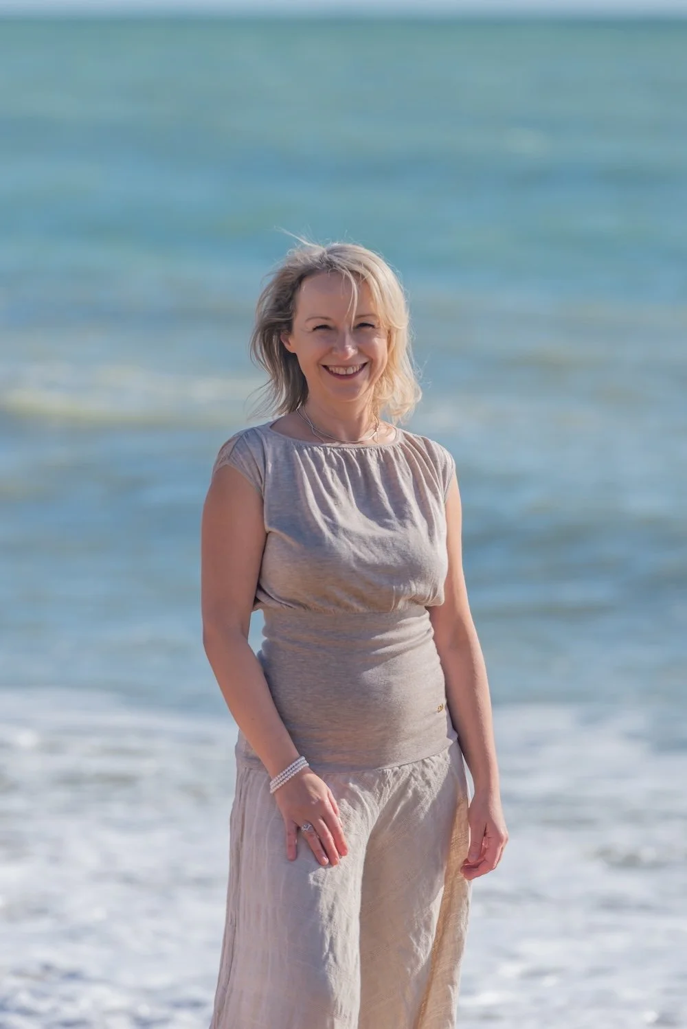 Smiling woman in a light dress stands on the beach with the blue sea and sky in the background.