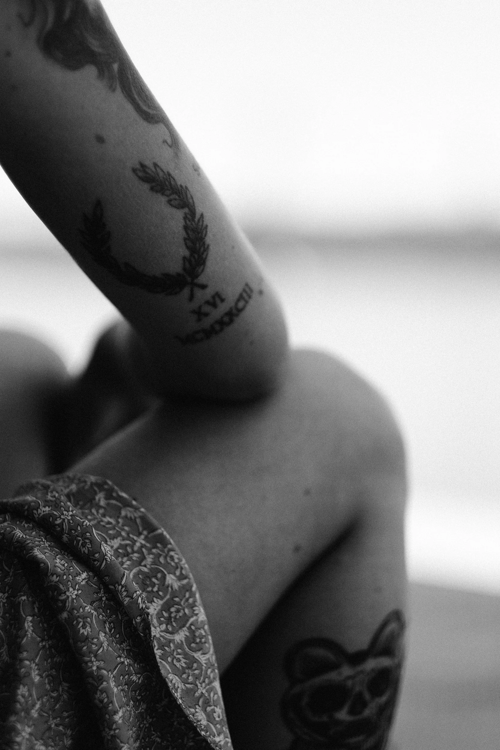 Close-up of a person's upper arm with tattoos, wearing a patterned garment, in black and white.