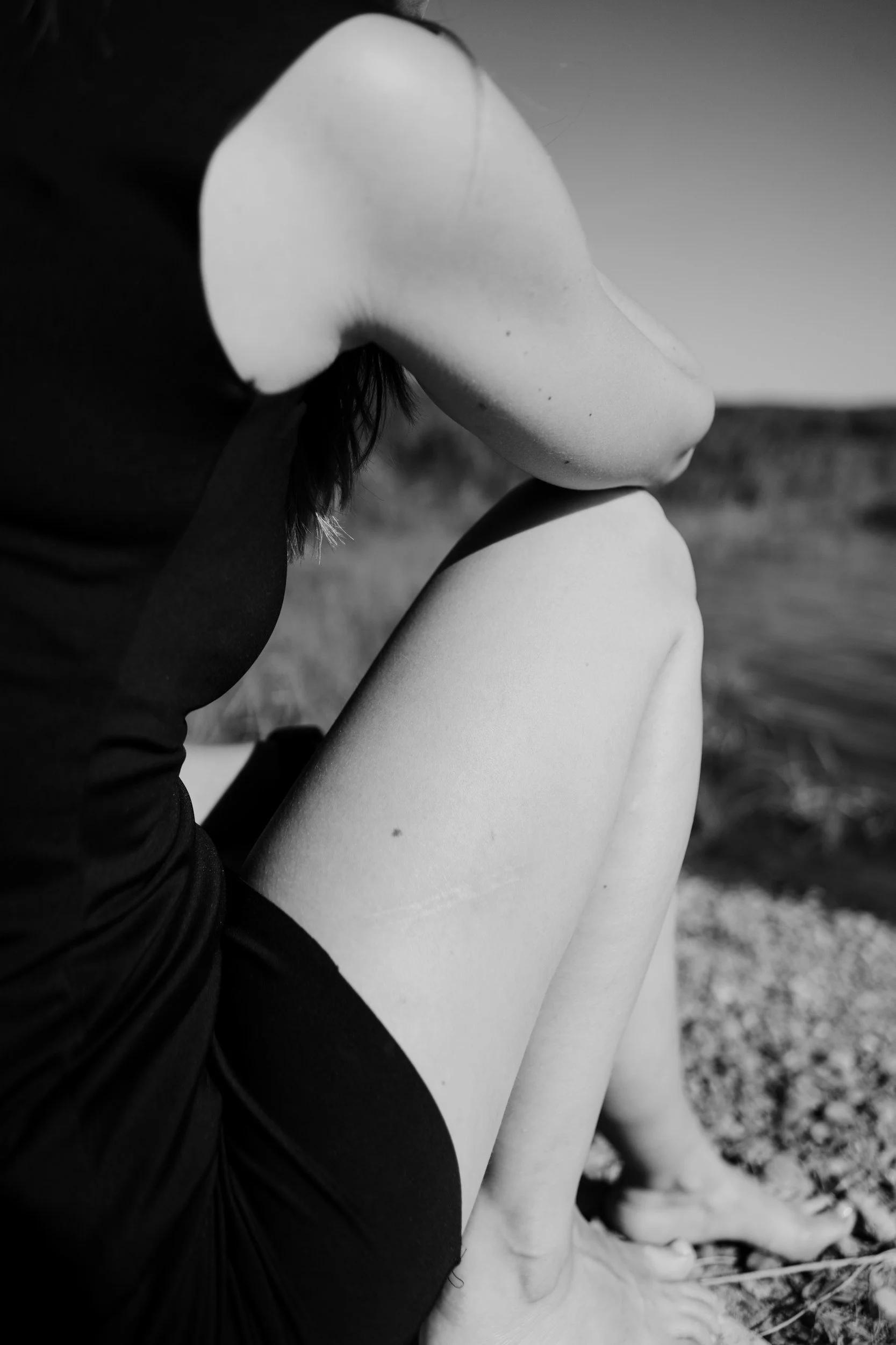 A black and white photo of a woman sitting on the ground outdoors, with her legs bent and arms crossed over her knees, wearing a dark shirt. She appears to have dark, tousled hair.