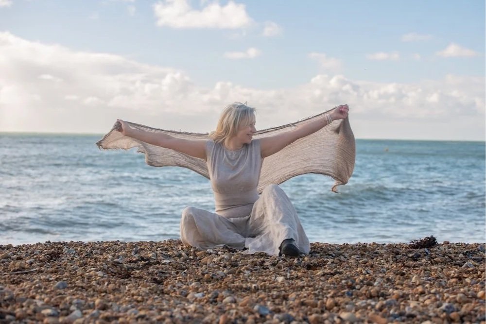 Woman sitting on pebbled beach near the ocean, holding a sheer scarf outstretched behind her with arms wide, smiling and enjoying the outdoors.