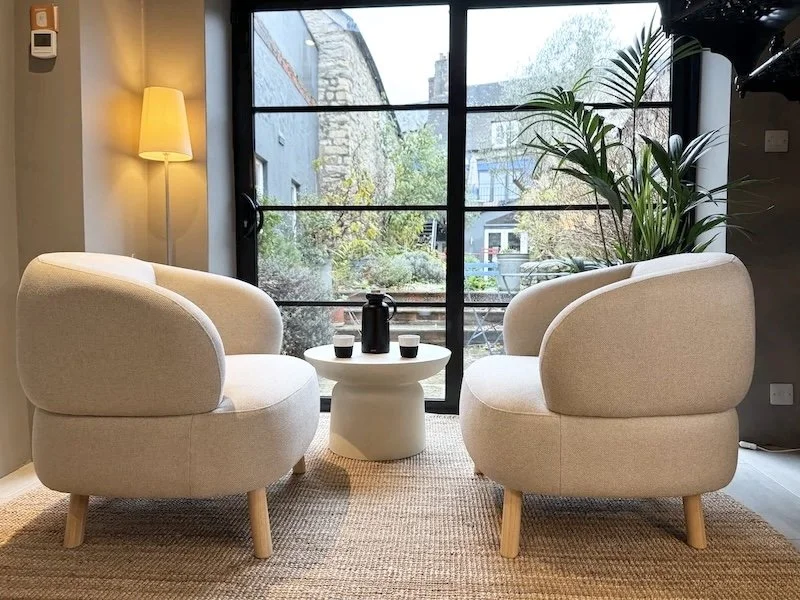 Cozy modern sitting area with two beige armchairs facing each other, a small round white coffee table with a black teapot and cups, a tall floor lamp, a large plant next to a large window overlooking a garden and a stone building outside.