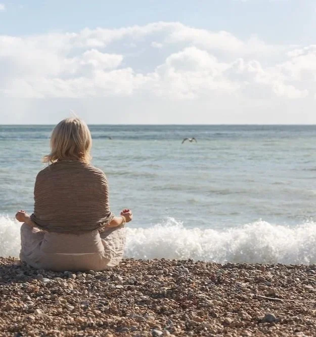 A woman with blonde hair sitting cross-legged on a pebbly beach, facing the ocean, with a bird flying over the waves in the distance.