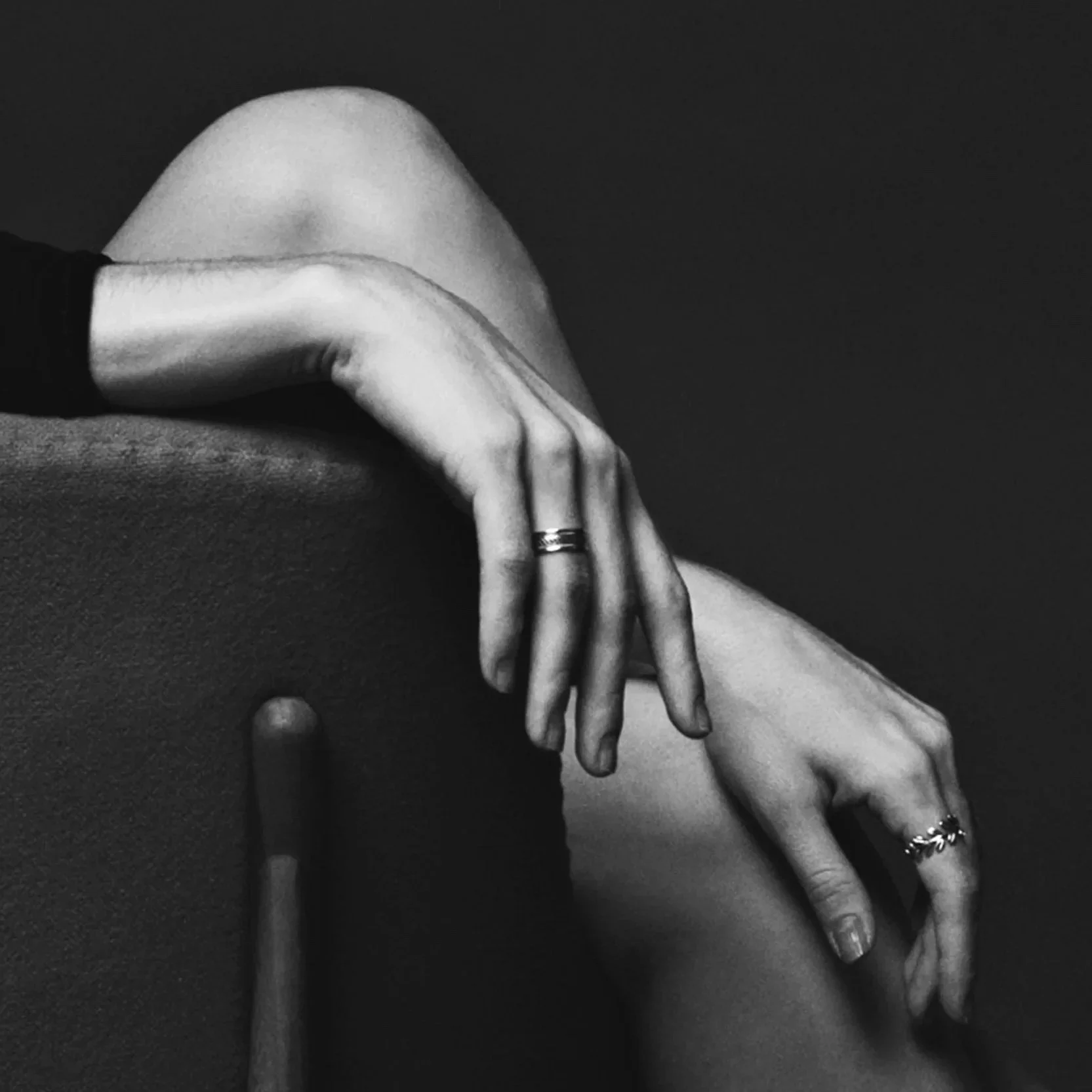Black and white photo of a person's arm resting on another person's lap with the hand visible, wearing rings. The background is dark.