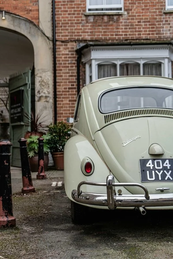 A vintage cream-colored Volkswagen Beetle parked in front of a brick building with a bay window and potted plants nearby.