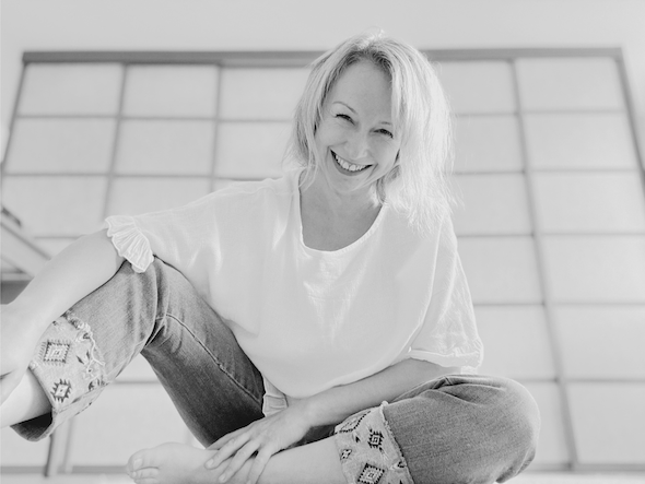A woman sits cross-legged, smiling warmly at the camera. She wears a light blouse and embroidered jeans in a bright room.