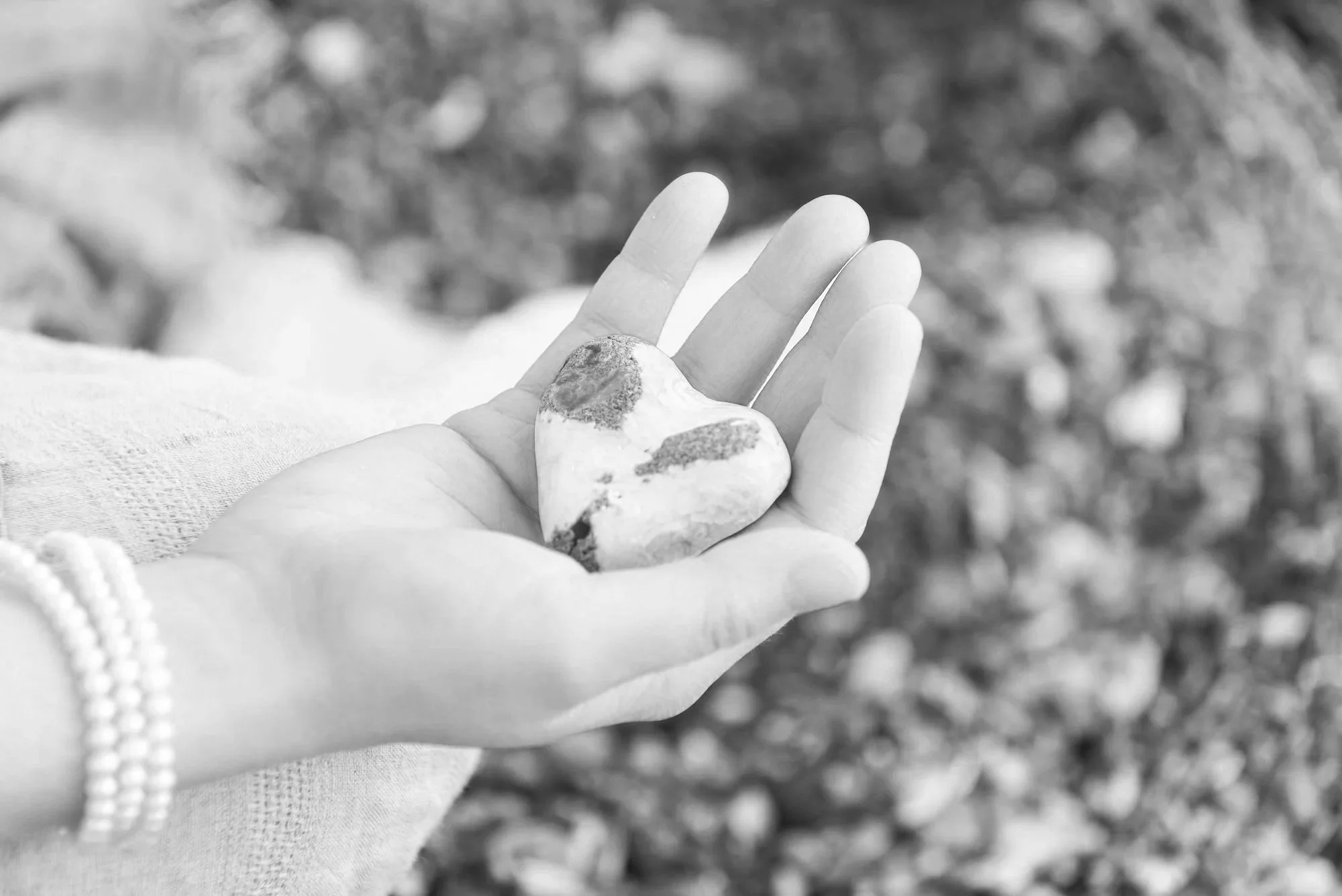 A hand holds a heart-shaped stone, with a blurred natural background and a pearl bracelet on the wrist.